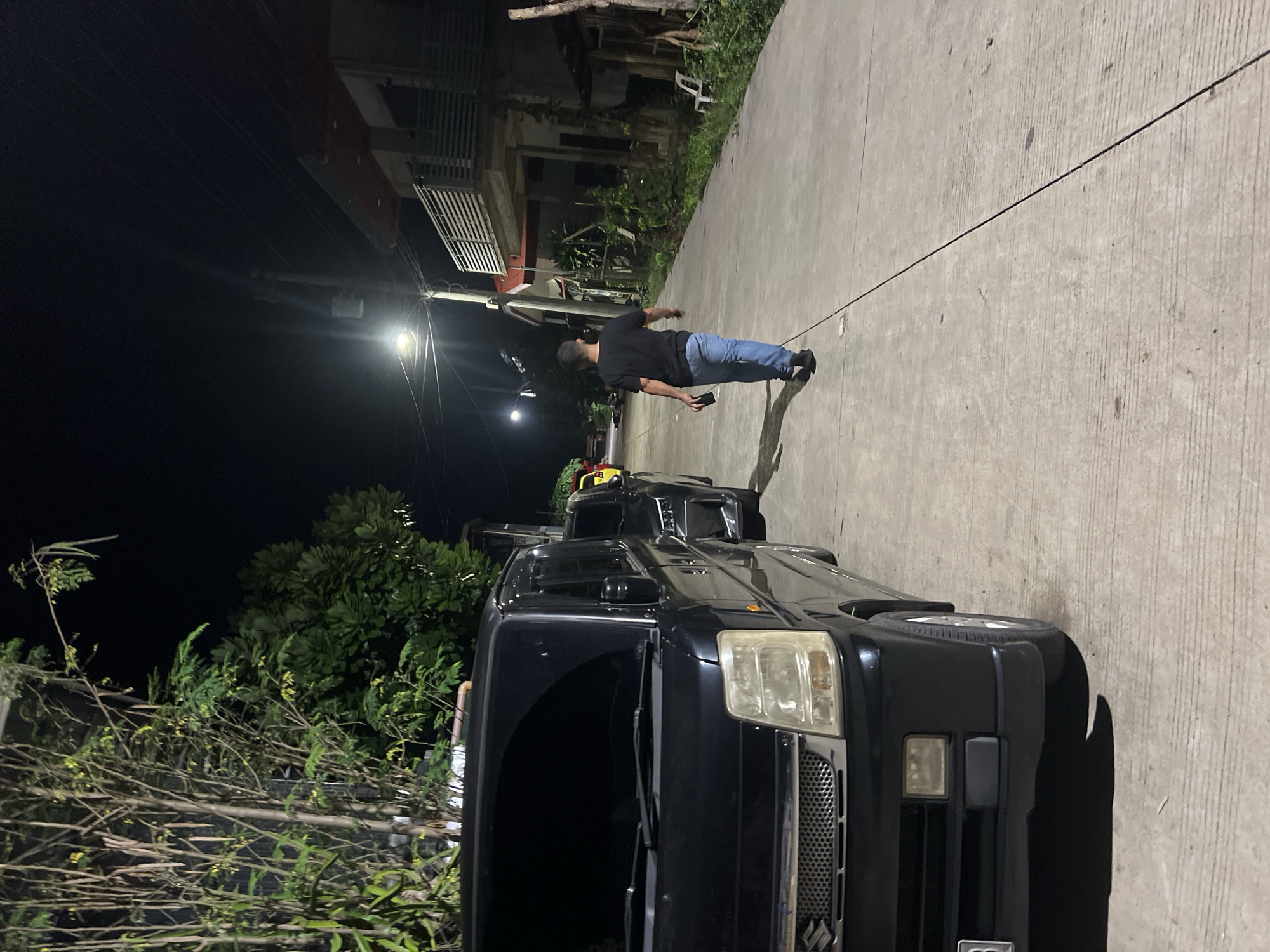 A black Suzuki car is parked on a paved road with a man standing nearby. The street is illuminated by a streetlight, and buildings are visible in the background. The scene is set in Davao City, Philippines. The man is dressed in a dark shirt and blue jeans and is looking at a cell phone in his hand. The car is facing the camera, revealing its front grill and headlights. Trees and foliage line the side of the road. The scene appears to be at night.