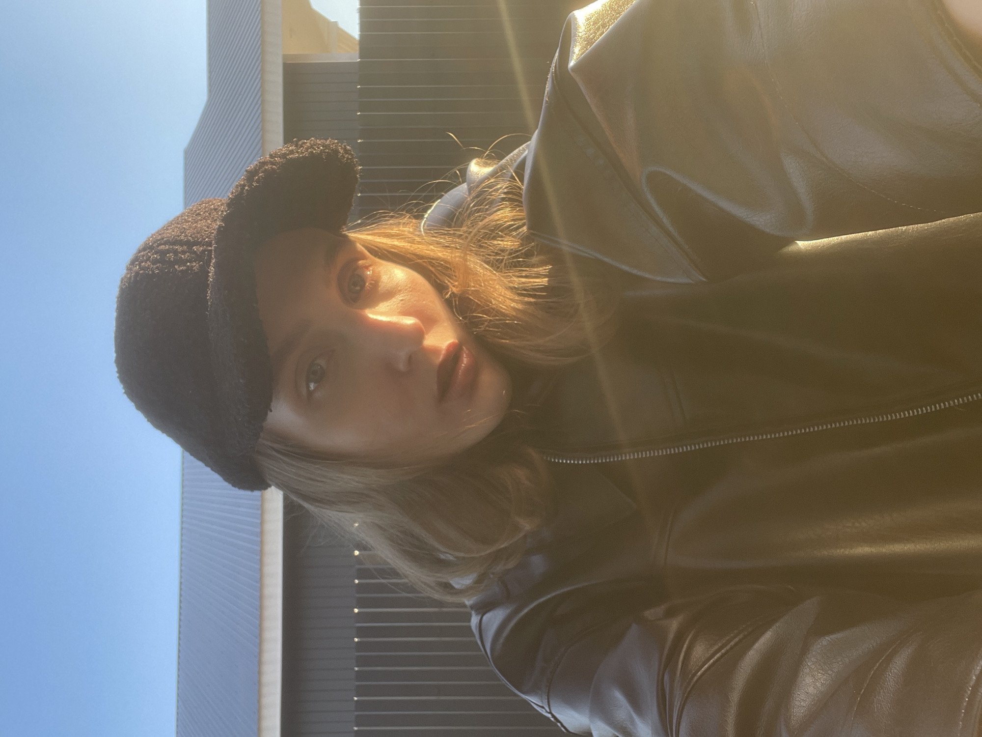A close-up portrait features a young woman looking directly at the camera, illuminated by strong, direct sunlight that creates significant lens flare. She has shoulder-length light brown hair, which catches the sunlight, and wears a dark, fuzzy baseball cap along with a dark, shiny jacket, likely leather or a similar material, with a visible zipper on the right side. Her expression is neutral.
The background on the left reveals a clear blue sky. On the right and behind her head, parts of modern buildings with distinct horizontal siding or panels are visible, suggesting an urban or semi-urban setting. The photo was taken outdoors during bright daylight under clear weather conditions. There is no visible text in the image. The location is Podshivalovo, Russia.