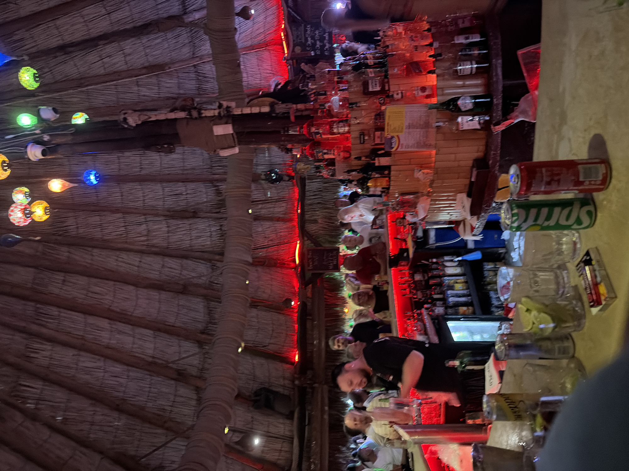 This image depicts the interior of a dimly lit bar, likely located in the city of Nesebar, Bulgaria, given the context. The bar has a rustic, thatched roof ceiling and is illuminated by red ambient lighting and colorful, decorative lamps hanging from above. A prominent feature is a bar counter laden with numerous bottles of alcohol. Behind the counter, a bartender, a male with dark hair and a beard, is visible, and a shelf stocked with more bottles is present. Several patrons are standing and seated at the bar, some with drinks in front of them. In the foreground, the bar top is visible with several glasses, cans of soda (Sprite and another red can), and a pack of cigarettes. The overall atmosphere appears lively and social, characteristic of a bar environment.
