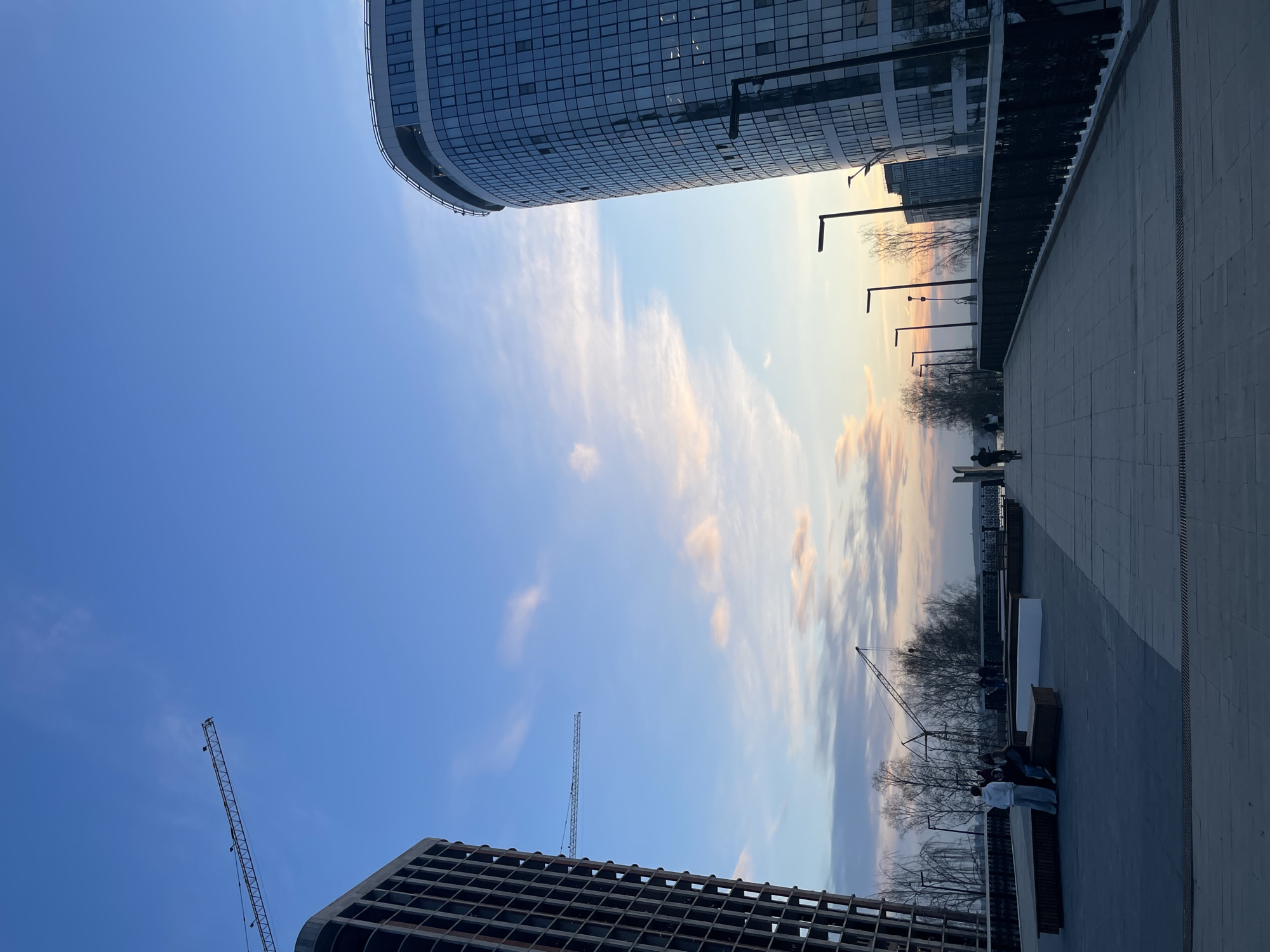The image captures an urban outdoor scene, likely a promenade or plaza, under a late afternoon or early evening sky. The clear sky transitions from a vibrant blue overhead to soft pastel hues of orange, pink, and yellow near the horizon, suggesting sunset. Wispy clouds are scattered across the warmer parts of the sky.
On the left, a tall building with an exposed, grid-like concrete and metal structure, possibly under construction, partially frames the scene. A large construction crane extends upwards from behind it into the blue sky. On the upper right, a modern, curved glass-fronted building stands prominently, reflecting the ambient light. A second, more distant construction crane is visible against the colorful horizon.
The right side of the image features a wide, light-colored paved walkway, detailed with drainage grates, stretching into the distance. It is lined with several contemporary, black L-shaped streetlights. Along the walkway, people are engaged in leisure activities; a few individuals are seated on wooden benches, while one person in a white top and light trousers stands facing away from the camera, looking towards the horizon. Beyond the path, a row of bare, deciduous trees suggests the time of year is late autumn or winter. A dark railing runs along the edge, hinting at a drop-off or an overlook onto a lower level or waterfront. The scene portrays a tranquil, developing urban environment. No visible text is present. This location is Izhevsk, Russia.
