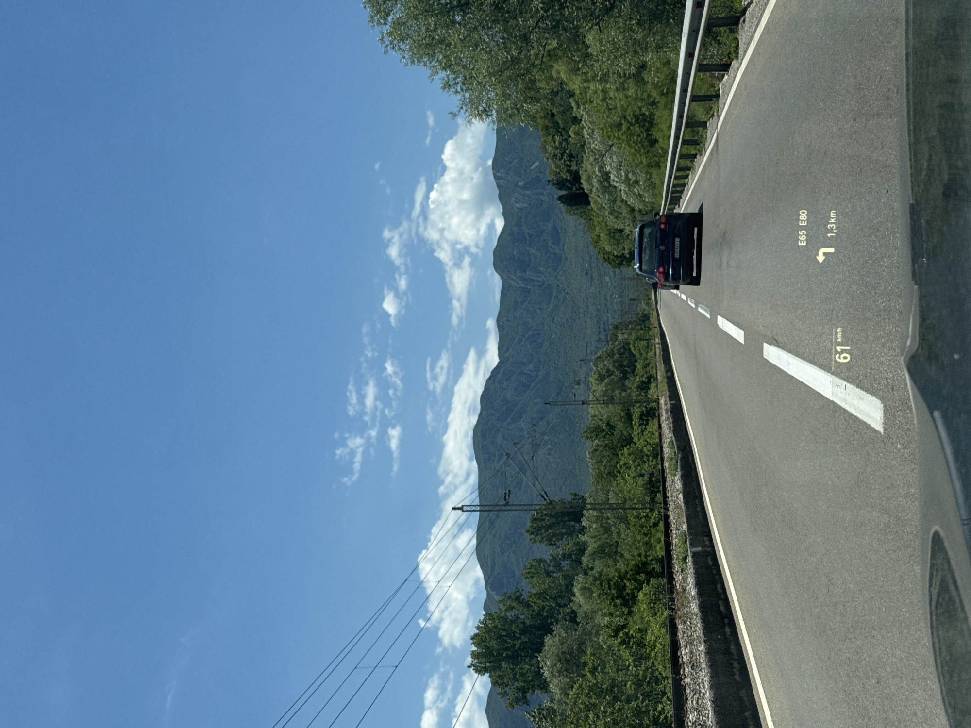 A black SUV is driving on a two-lane asphalt road through a mountainous, green landscape. The road has a white dashed line down the center and a white line on the right edge. There is a guardrail on the right side of the road. In the distance, there are green mountains under a blue sky with scattered white clouds. Overhead power lines are visible on the left side of the road, running parallel to a set of train tracks. A heads-up display on the windshield shows the speed as 61 km/h, a left turn instruction for E65 E80 in 1.3 km. The scene appears to be on the E65 road near Virpazar, Montenegro.