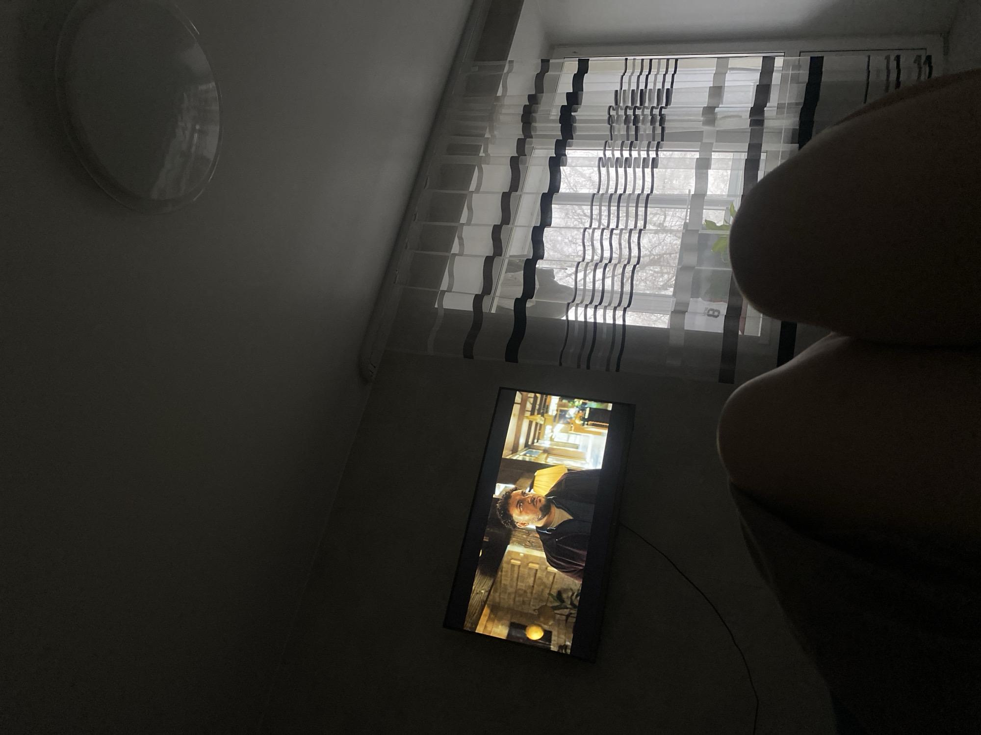A dimly lit interior shot with a partial view of a person's body on the right side of the frame. A window covered with striped blinds is above a TV, which is playing a scene with a man in it. A round light fixture is on the wall to the left. The location is Chaikovsky, Russia.