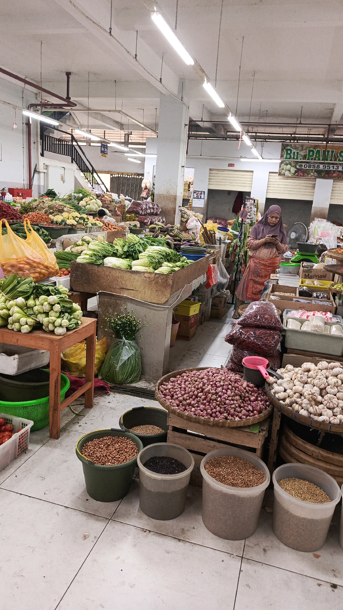 The image depicts an indoor market scene in Nologaten, Indonesia. The market is filled with various fresh produce and dry goods displayed on stalls and in containers. In the foreground, several large buckets contain different types of grains and legumes, including peanuts, black seeds, and lighter colored seeds. A basket overflowing with shallots is visible to the right of these buckets, next to another basket of garlic. Behind these items, a woman wearing a hijab and a colorful skirt is looking at her phone while standing near a stall. The stall itself features a large quantity of dried red chilies in plastic bags, as well as other items. Further back, more produce such as cabbage, lettuce, and other vegetables are arranged on tables. The market appears to be well-lit with fluorescent lights and has a concrete floor. The background shows more stalls, shelving units, and part of a staircase. The overall impression is a busy, well-stocked local market.
