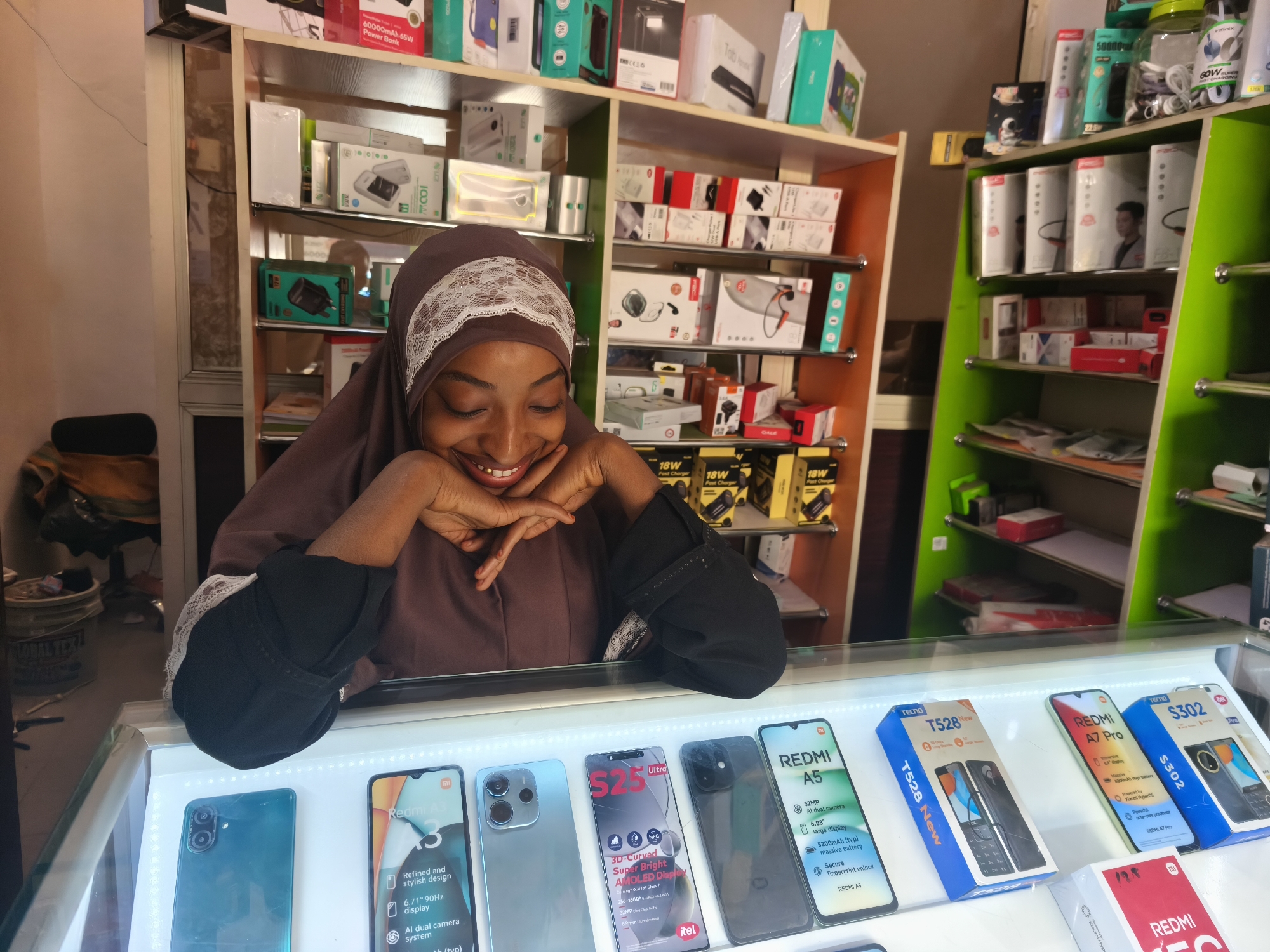 A young woman wearing a brown hijab and black sleeves is behind a glass counter in what appears to be a mobile phone shop. She rests her chin on her hands and smiles at the display of smartphones in front of her. The phones are mostly Redmi and Tecno brands, with a broken Itel S25 Ultra also present. Behind her are shelves stocked with boxes of accessories, including chargers, power banks, and headphones. The shelving is a mix of light brown/orange and green. The setting appears to be a commercial space in Jos, Nigeria, based on the brands of mobile phones.
