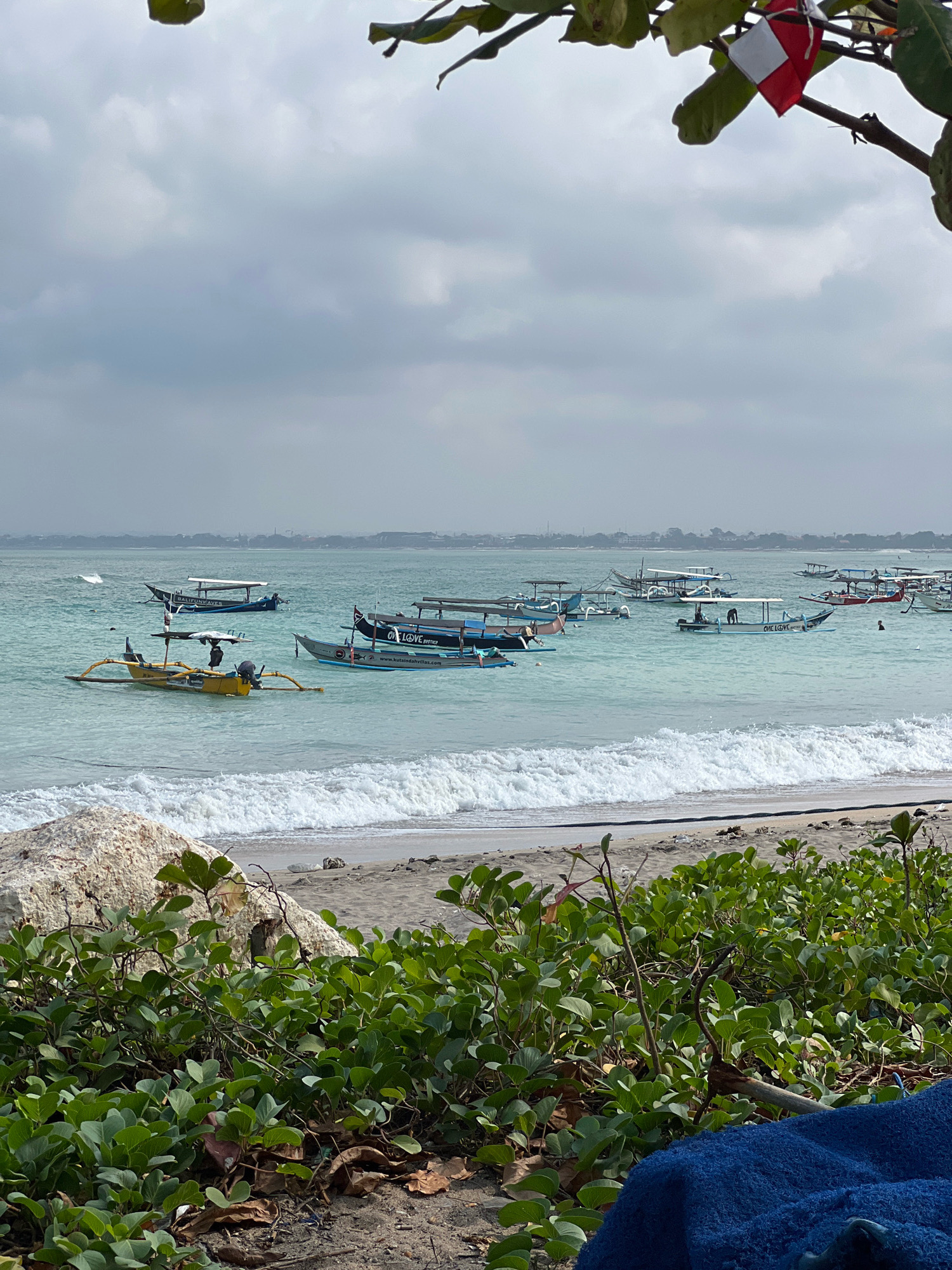 The image depicts a coastal scene in Kuta, Indonesia. Numerous traditional Balinese outrigger boats, known as jukungs, are anchored in the shallow, turquoise waters of the Indian Ocean. The boats vary in color, with some being yellow, blue, and black, and many feature canopies providing shade. Several individuals are visible in and around the boats, either standing, sitting, or swimming. The foreground of the image shows a sandy beach with green vegetation, including morning glory vines, and a large rock. In the background, the hazy outline of the coastline and buildings of Kuta is discernible. The sky is overcast with diffused light. A small red and white flag is attached to a branch of a tree in the upper right corner. The overall impression is one of a typical day at a busy beach in Kuta, with local fishing and tour boats present.