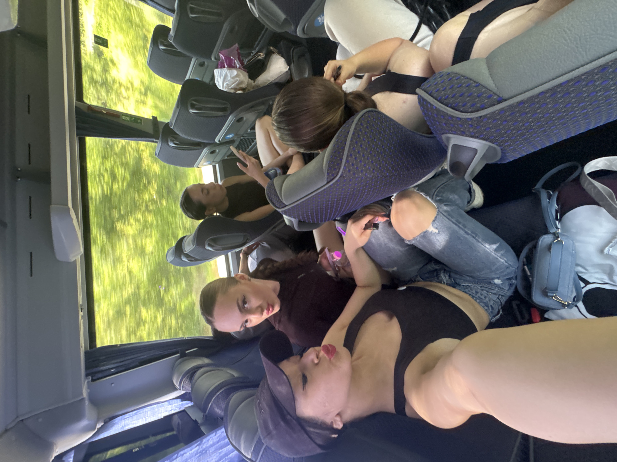 The image captures the interior of a moving passenger coach bus during daylight hours. The scene is located in the city of Nikolaevo, Bulgaria. Four young adult women are visible, occupying multiple seats and the aisle space.
In the immediate foreground, a woman wearing a black baseball cap, black tank top, and ripped blue jeans is taking a selfie. She has dark hair, prominent red lipstick, and is making a puckered lip expression towards the camera. Her right arm is extended, holding the camera, and her left arm rests on her bent knee. A light blue shoulder bag with a gold chain strap rests on the floor near her left leg.
To her right and slightly behind, a second woman, wearing a dark reddish-brown long-sleeved top and ripped blue jeans, looks directly at the camera with a slight smirk. Her dark hair is pulled back, and she holds a clear plastic bottle with a pink cap in her right hand.
Further back, seated by a window, a third woman with dark hair pulled back in a bun or ponytail is engrossed in her smartphone, looking down at the screen. She wears a black sleeveless top and has bare legs crossed at the ankles.
Partially visible in the background, behind a seat, a fourth woman with dark hair in a low ponytail or bun is seen with her hand near her ear, holding a dark pen-like object. She also wears a black top.
The bus interior features dark blue patterned seating with grey trim, grey overhead storage compartments, and large windows revealing blurred green foliage, indicative of travel through a vegetated area. Blue patterned curtains are partly drawn on the left side of the bus. A white cloth or plastic bag is placed on a seat in the far right background.