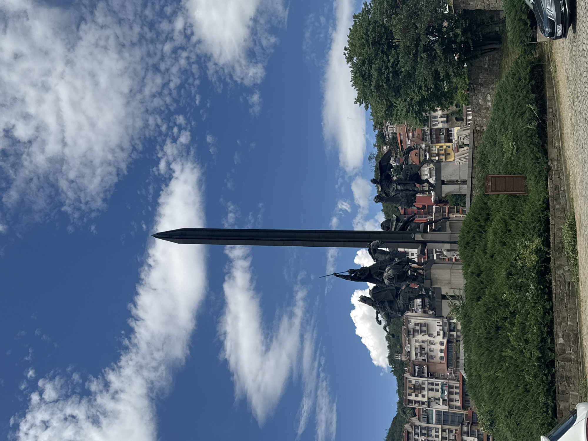 The image shows a statue of horsemen with a tall, dark obelisk behind them, situated on a hill in Veliko Tarnovo, Bulgaria. The statue depicts several figures on horseback, some with weapons, in a dynamic pose. The obelisk is slender and pointed, reaching towards a bright blue sky filled with white, fluffy clouds. Below the statue, there is a neatly trimmed green hedge. In the background, the city of Veliko Tarnovo unfolds, with its characteristic colorful buildings clinging to the hillside. The architecture appears traditional, with terracotta roofs and multi-story structures. A large, leafy green tree is visible on the right side of the image, partially obscuring some of the buildings. In the foreground, there is a stone wall and a paved area with cobblestones. A dark-colored car is parked on the right, with its license plate visible. The overall scene suggests a sunny day in a historic city.
