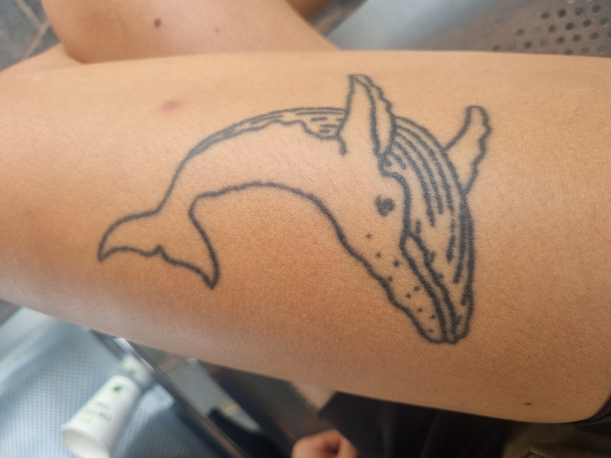 A close-up shot shows a person's arm with a black ink tattoo of a humpback whale. The tattoo depicts the whale in a curved posture, with details like its fins, tail, and characteristic throat grooves visible. The arm is tan in color, and some blemishes are present on the skin. In the lower left corner of the image, the edge of a white bottle with green text is visible, resting on a metallic surface with perforations. The arm appears to be resting on a lap covered in black material. The image does not contain any specific markers or identifiers relating it to the city of Bangaliguda, India.