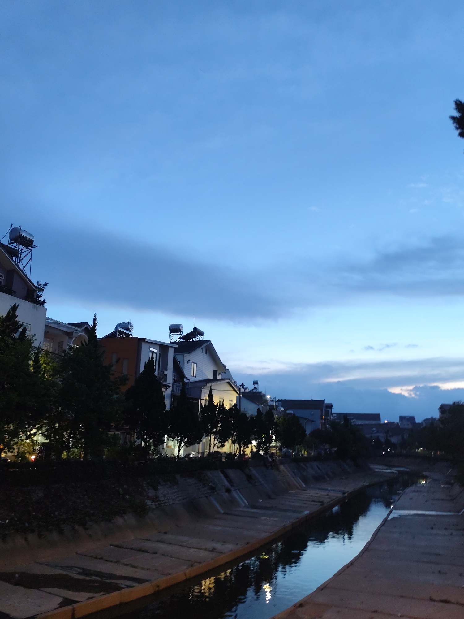 This image captures a tranquil evening scene in Da Lat, Vietnam, along what appears to be a concrete-lined canal or waterway. The sky above is a deep blue, indicative of dusk or early night, with scattered clouds that show varying shades of grey and lighter blue towards the horizon.
Along the banks of the canal, a row of residential buildings stands, featuring multiple stories and diverse architectural styles. Many of these houses have prominent solar water heaters installed on their rooftops, suggesting a focus on sustainable energy. Lights are visible in some windows, casting warm yellow glows that contrast with the cooler tones of the sky and water. Dense green trees line the canal's edge, partially obscuring the lower portions of the buildings and creating a sense of natural integration within the urban setting.
The canal itself is narrow and appears to be man-made, with concrete slopes leading down to the water. The water is calm and dark, acting as a mirror that reflects the blue sky and the artificial lights from the houses, creating shimmering streaks on its surface. No people or specific activities are visible, contributing to the peaceful and quiet atmosphere of the scene. The overall impression is one of a serene urban landscape winding down for the night.