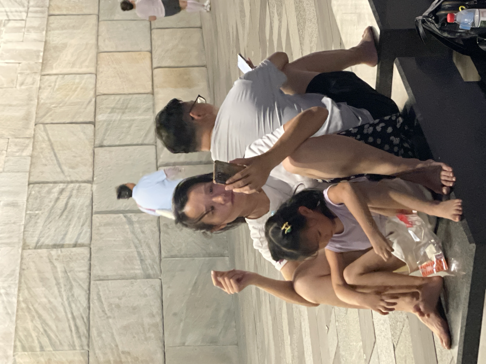 In Yuzhong District, China, two adults and a child are seated on a stone bench against a backdrop of large, light-colored stone blocks. The adult female in the foreground is holding a mobile phone and appears to be showing it to the child. The child, a young girl, is looking down at something in a clear plastic bag near her feet. Beside them, an adult male, wearing glasses, is also seated and looking at his own mobile phone. Further in the background, two other individuals are visible walking away from the camera. A black backpack and a water bottle are on the ground to the right of the seated group. The ground is paved with light-colored tiles.