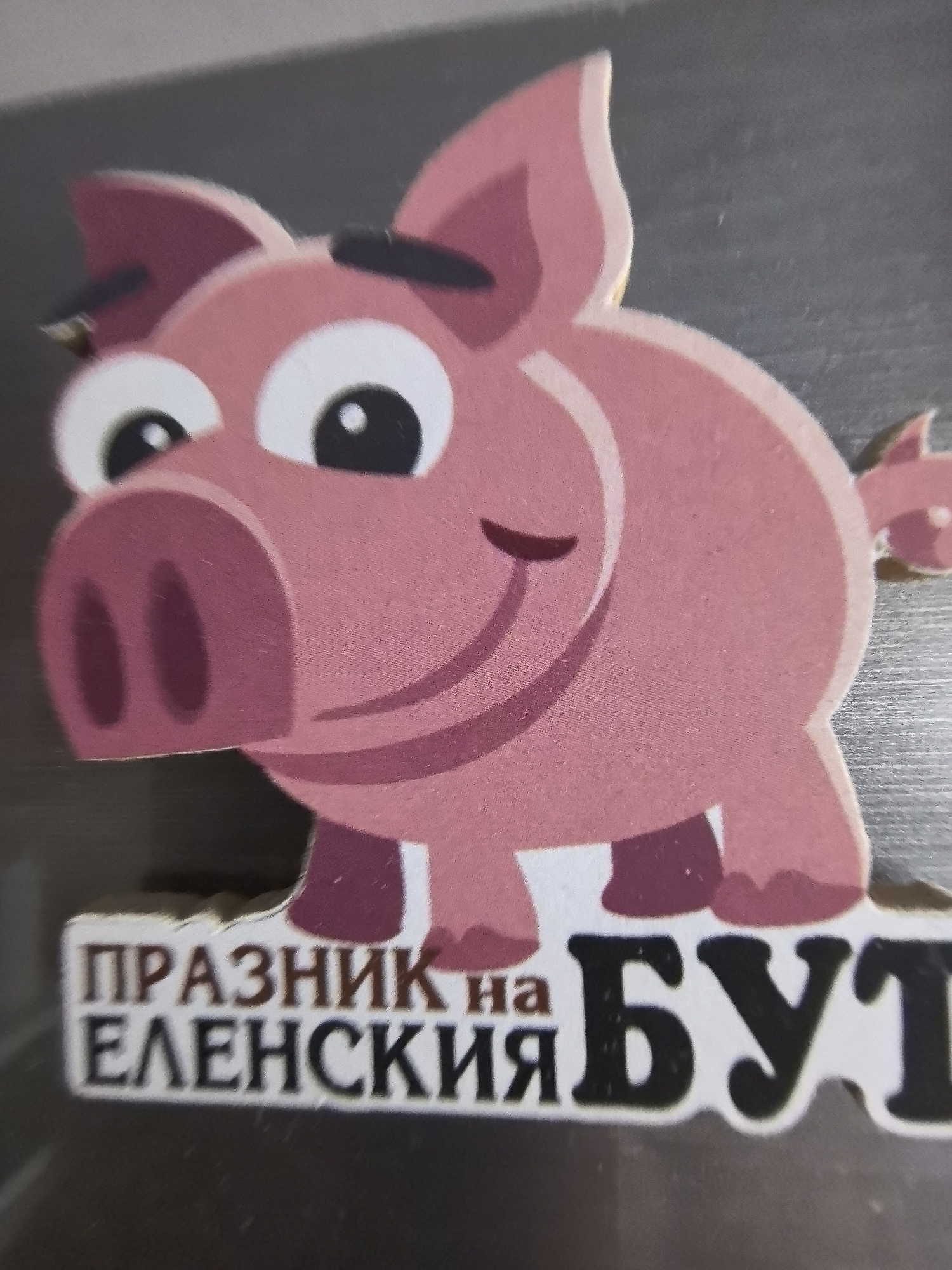 The image shows a sticker or magnet with a cartoon pig on it. Below the pig, there is text in Cyrillic, which translates to "Holiday of Elena and But". Elena and But are two small settlements in the Burgas province of Bulgaria. The background is a flat, possibly metallic, grey surface. No people are visible in the image.