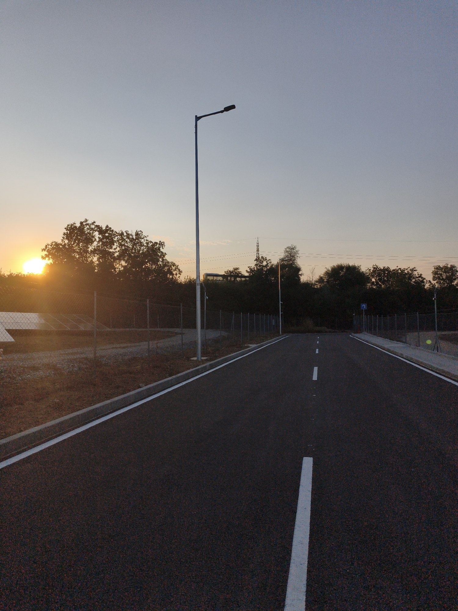 The image depicts an asphalt road with white lane markings, leading into the distance. On the left side of the road, a chain-link fence encloses an area with solar panels and vegetation. A tall lamppost stands beside the road, and a distant bus can be seen. The sun is setting behind silhouetted trees, casting a warm glow across the sky. The scene is consistent with an industrial or semi-rural area. The location is identified as Lovech, Bulgaria.