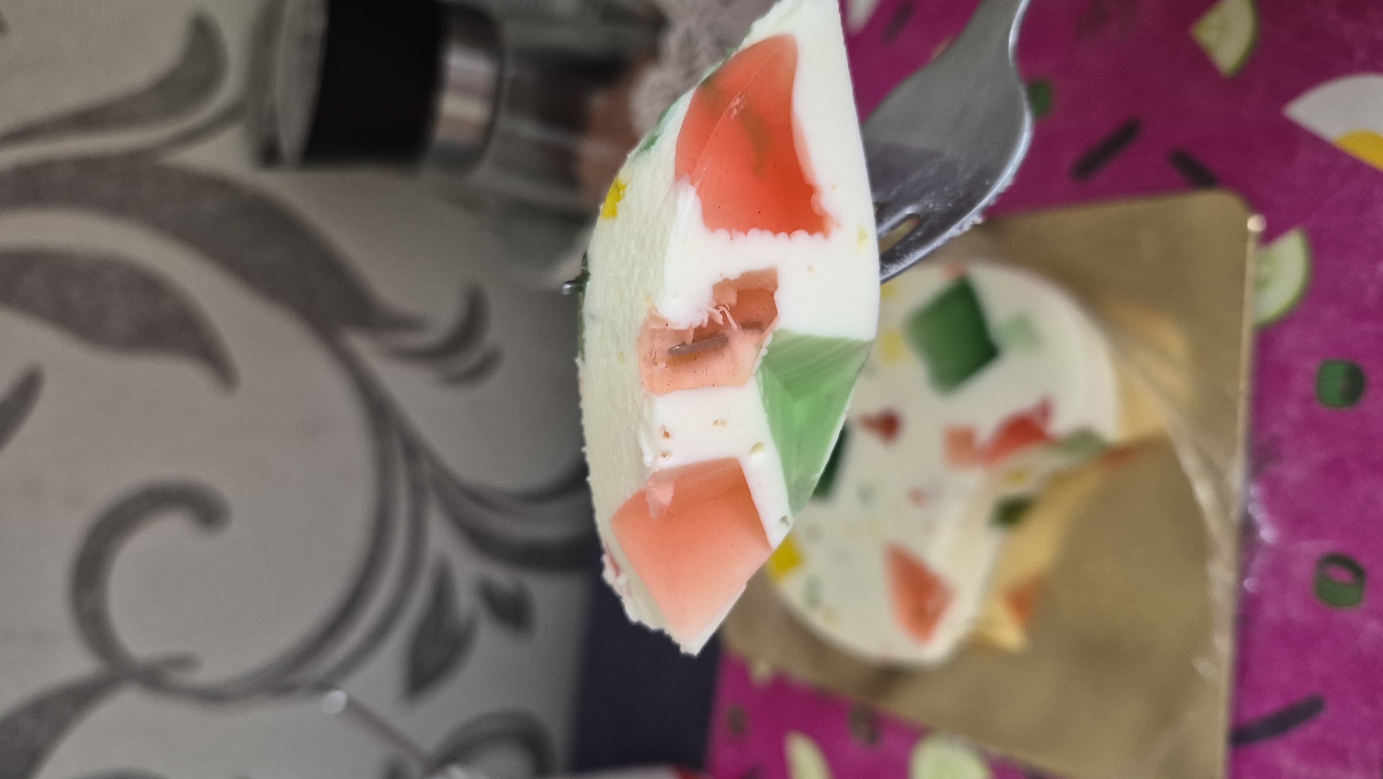 A slice of colorful layered jelly dessert is held on a fork in the foreground, against a blurred background. The dessert has distinct layers of white, pink, green, and yellow gelatinous material, forming geometric shapes. A larger, round jelly cake with the same colorful inclusions is visible behind the slice, resting on a golden plate. The background features a textured wall with a subtle floral pattern and a glass container with a dark lid. The scene appears to be indoors, possibly in a kitchen or dining area. The image does not contain any information related to the city of Krauja, Latvia.