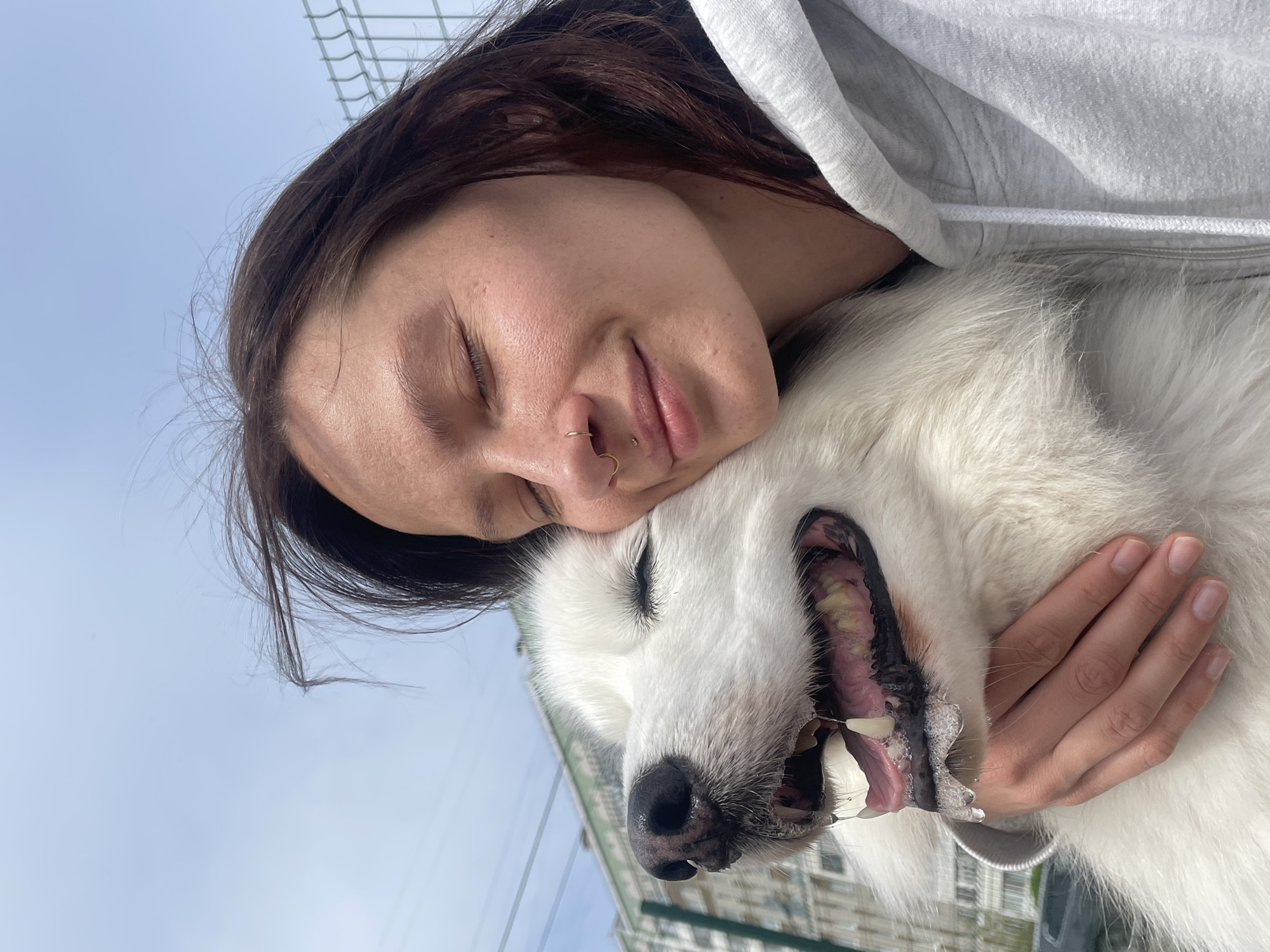 A woman with long brown hair is embracing a large white Samoyed dog outdoors. The woman is wearing a light gray hooded sweatshirt. Her eyes are closed, and she has a nose ring. The dog's mouth is open, exposing its teeth and tongue. The background includes a wire fence and buildings. The location is Izhevsk, Russia.