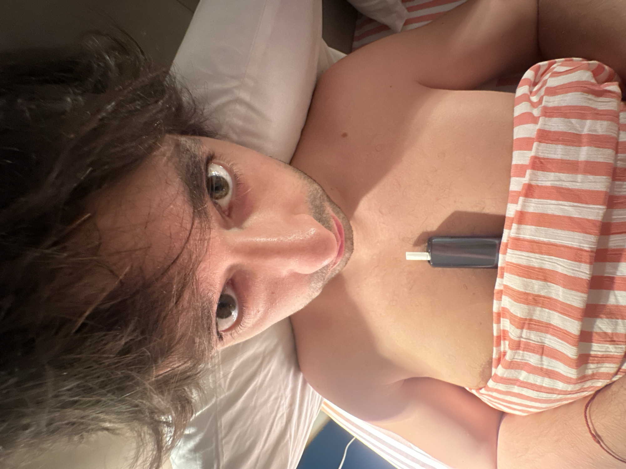 The media file is a close-up, overhead shot of a person lying in bed, with their head tilted back and eyes wide open. The person has dark, messy hair and is looking directly at the camera. They are shirtless, and a pink and white striped blanket is draped over their lower torso. A small, dark cylindrical object with a white tip is resting on their chest, partially covered by the blanket. The setting appears to be a bedroom, with a white pillow visible behind the person's head. The lighting suggests it might be nighttime or an indoor setting with artificial light. The person's expression could be interpreted as surprised or concerned. No visible text is present in the image. The context provided indicates the location is Moscow, Russia.