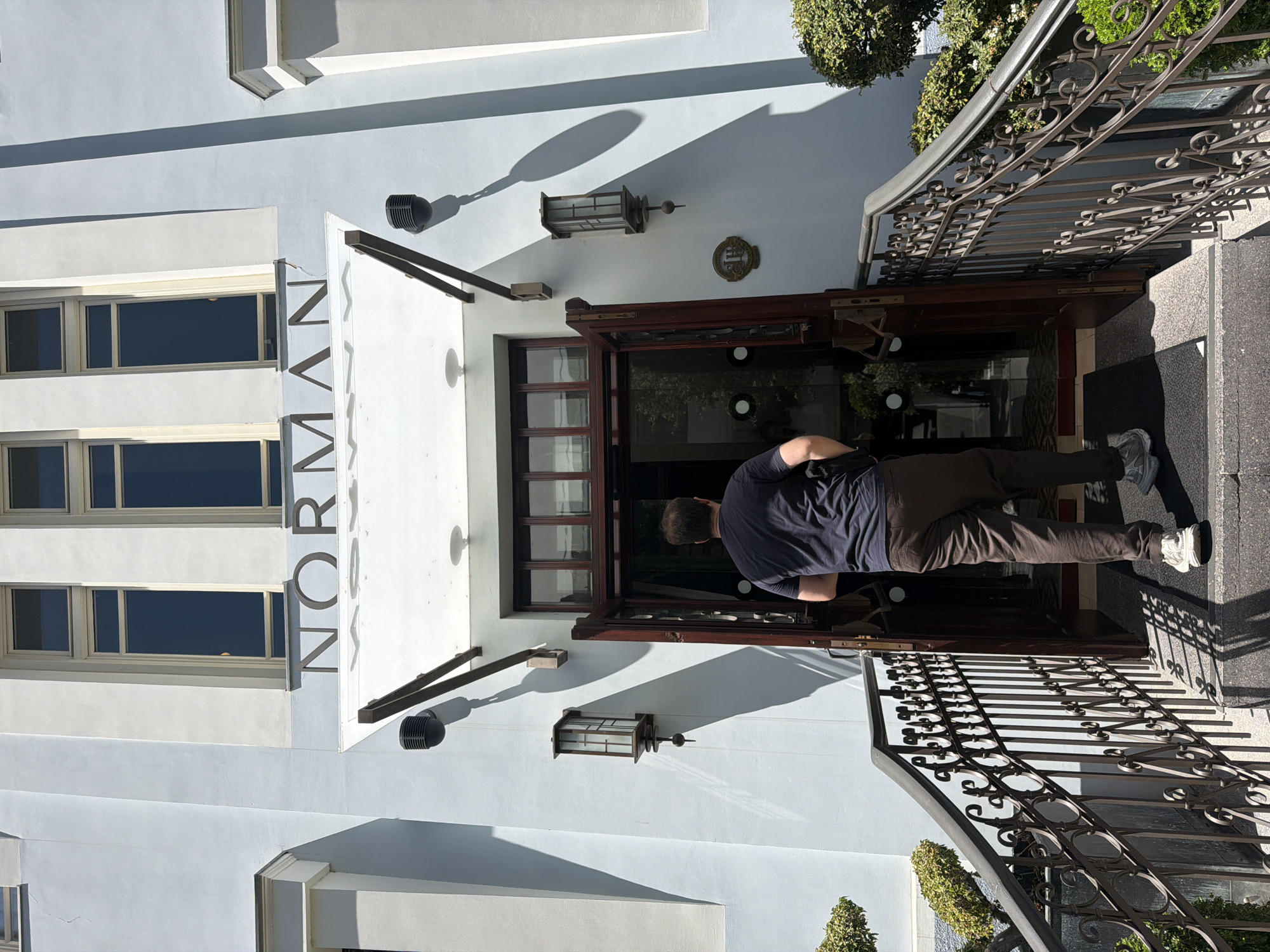 A person wearing a dark blue t-shirt and brown pants walks up a set of stairs towards the open entrance of a building. The building's facade is painted a light blue, with three tall, narrow windows visible above the entrance. The word "NORMAN" is inscribed in large, dark letters above the entrance, which is sheltered by a white awning. Two ornate lamps are mounted on the building's exterior walls, flanking the entrance. Metal railings with decorative scrollwork line the sides of the stairs. Green bushes are planted on either side of the entrance. The scene appears to be in Tel Aviv, Israel, given the architectural style and the name "Norman" potentially referring to Norman’s, a hotel and restaurant located in the city.