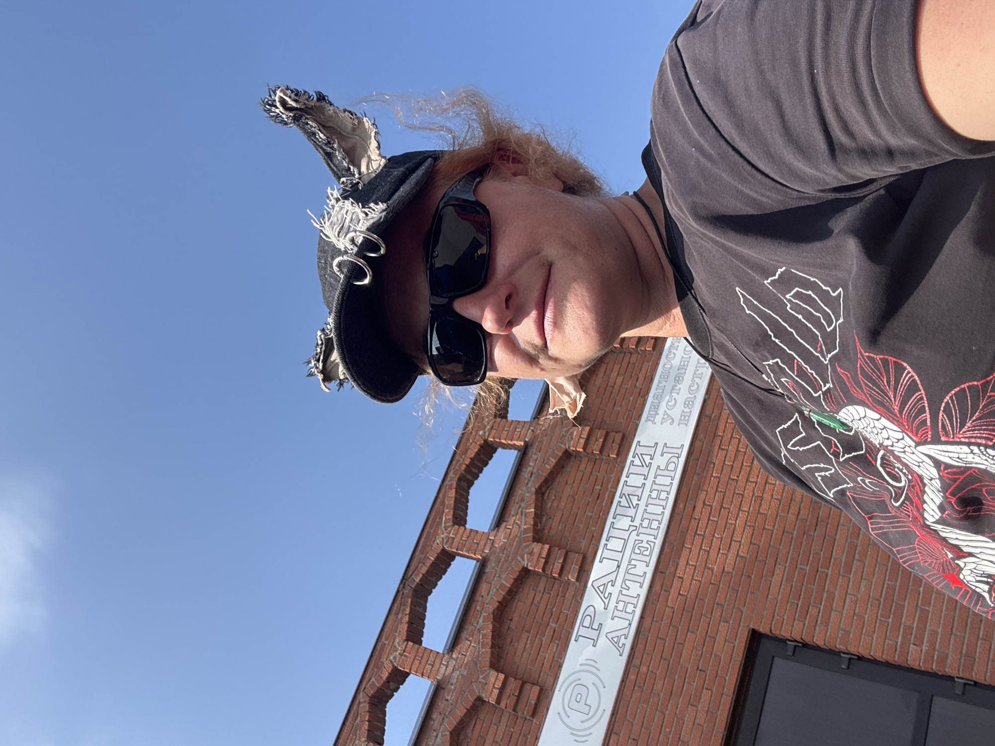 A person with reddish hair, wearing sunglasses and a black cap with torn denim "ears" adorned with metal rings, is pictured outdoors against a clear blue sky. The person is wearing a dark t-shirt with a red and white graphic design. Behind the person, a brick building with arched window openings is visible. A sign on the building reads "РАЦИИ АНТЕННЫ" which translates to "RADIOS ANTENNAS" in English. The location is identified as Verkh-Sosnova, Russia. The lighting suggests it is daytime.