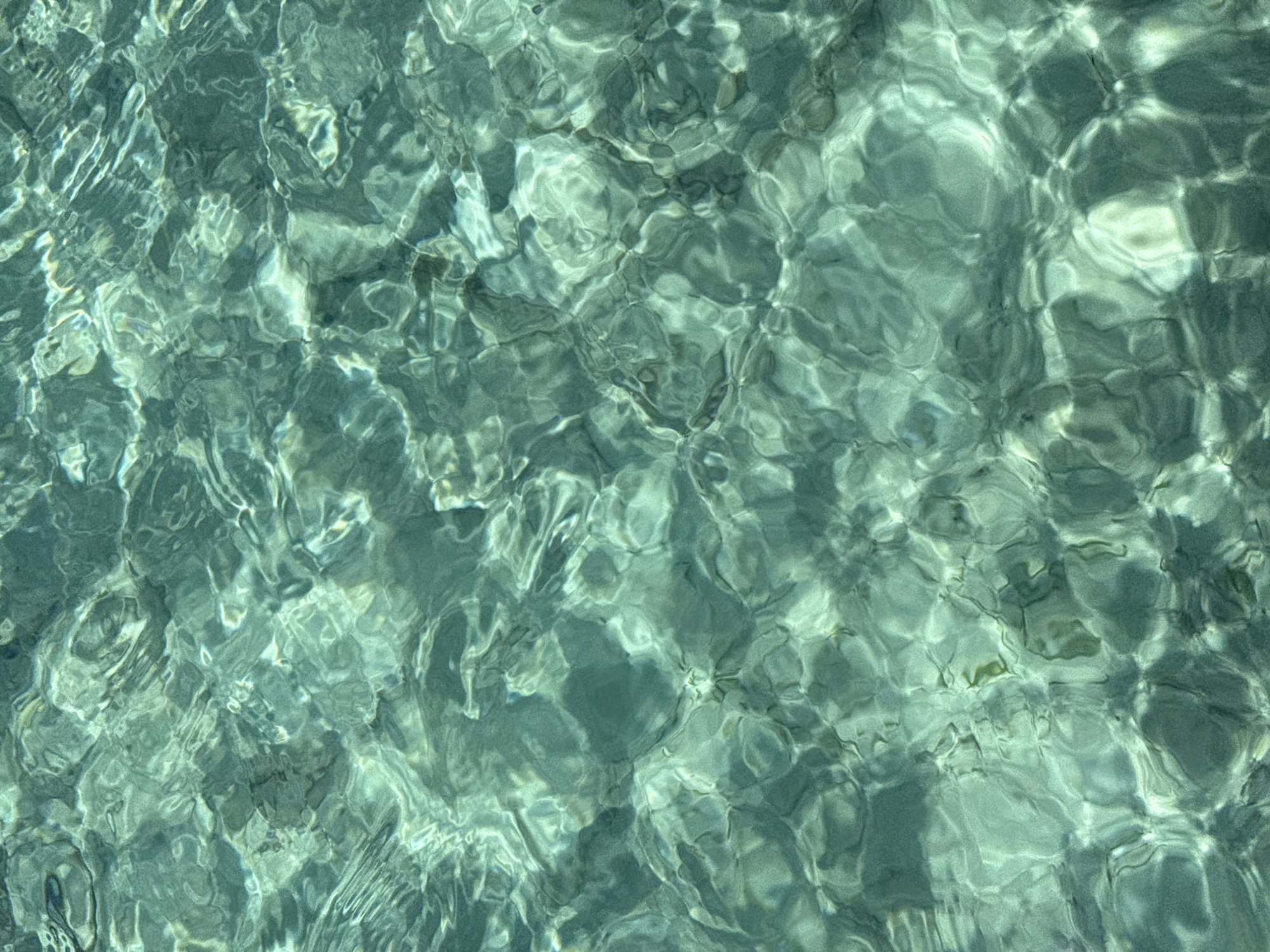 The image depicts a close-up view of shallow water. The water is clear and slightly greenish, with sunlight refracting through the surface, creating patterns of light and shadow on the bottom. The location in the city of Unknown, Indonesia is based on the assumption that the image was taken in a coastal area in Indonesia, which is known for its tropical waters.