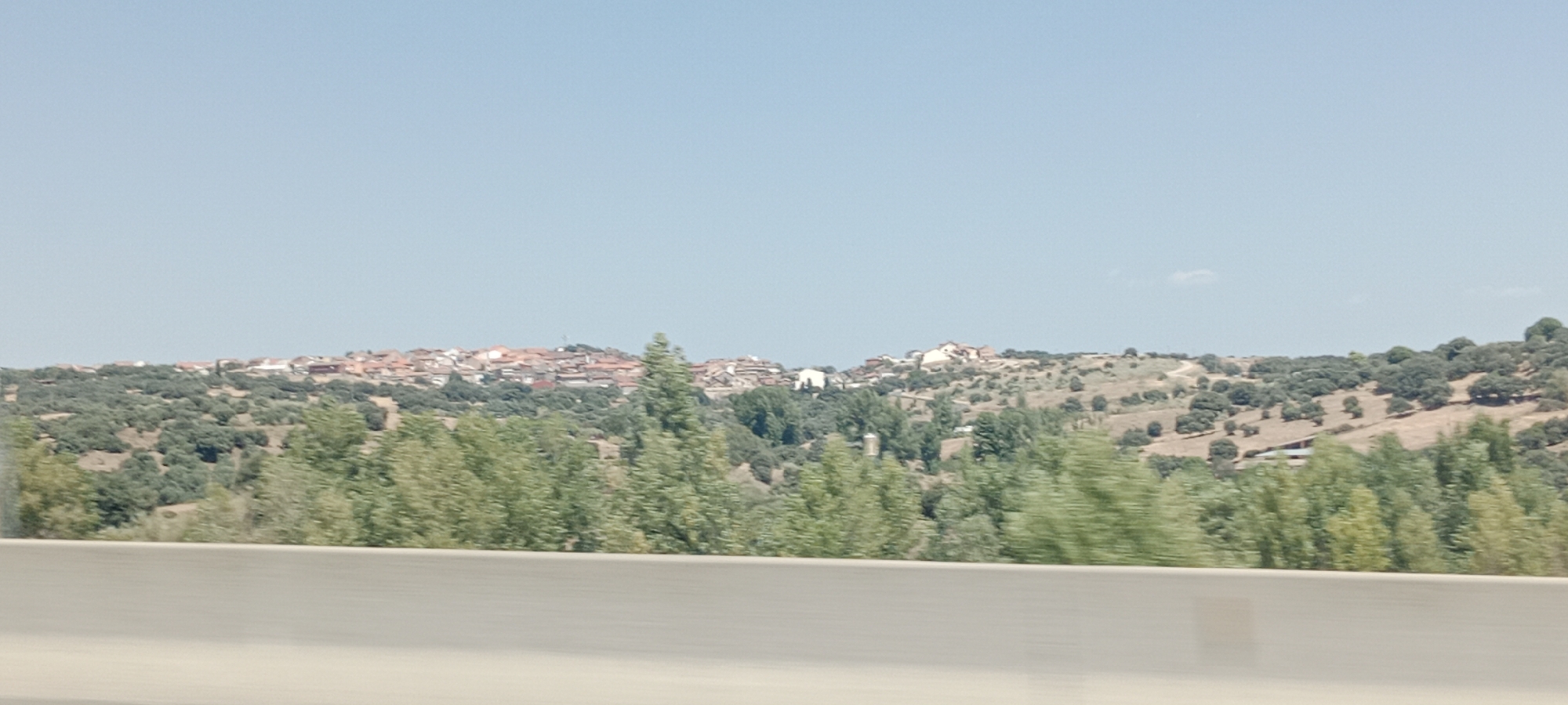 The image depicts a panoramic view of a town situated on a hillside, likely El Vellón, Spain, under a clear, pale blue sky. The town is characterized by a cluster of buildings with red-tiled roofs, nestled amidst rolling, arid hills covered in sparse vegetation and scattered trees. In the foreground, a blurred concrete barrier suggests a roadside perspective, possibly from a vehicle. The middle ground is dominated by a dense line of trees, with some appearing to be in motion, indicating a sense of movement within the frame. The overall impression is a sun-drenched, rural landscape with a village perched on a distant ridge.