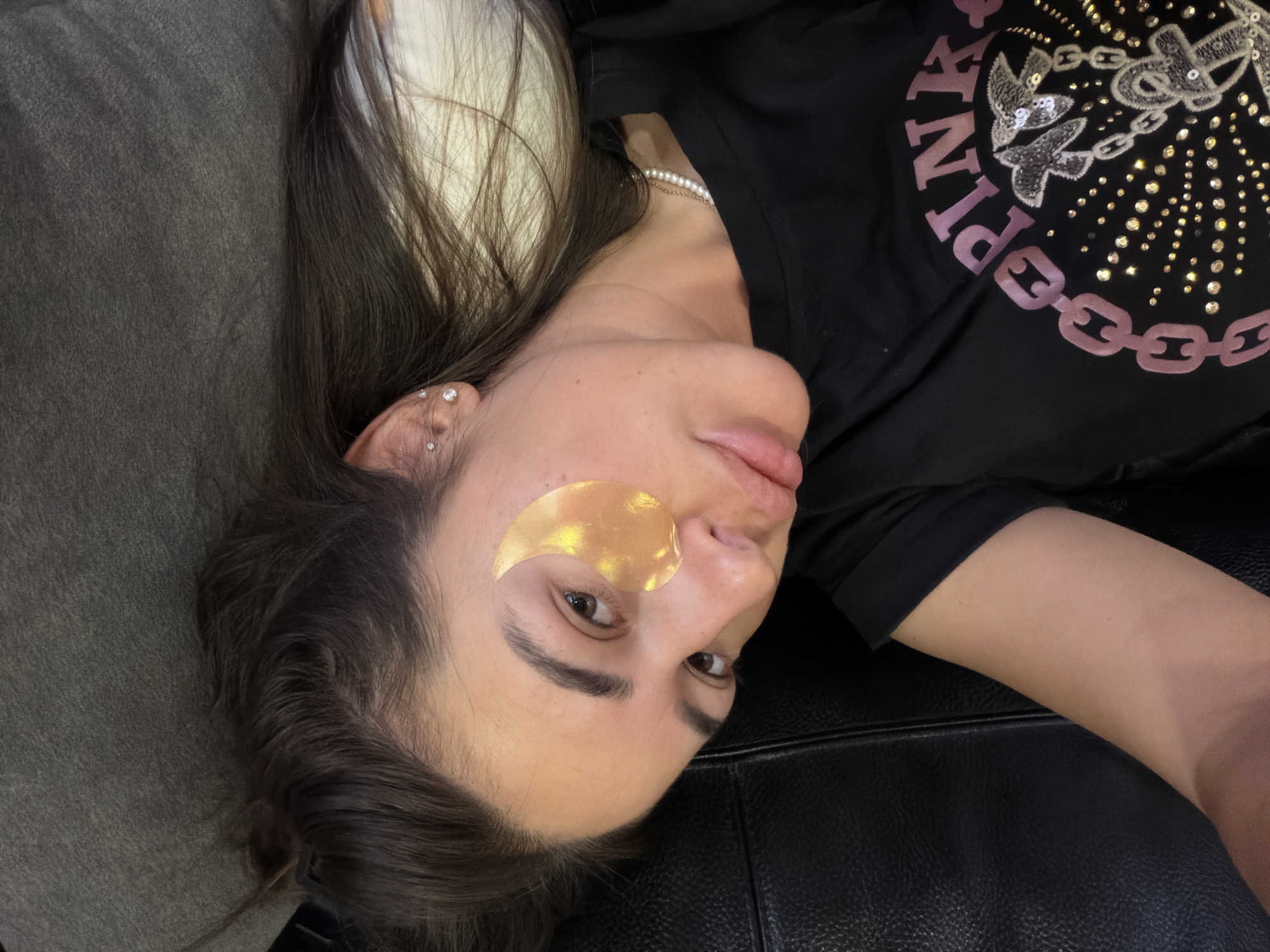 A woman is lying down with a skincare patch under her eye, and she is wearing a black t-shirt. The environment appears to be a bedroom in Novi Iskar, Bulgaria.