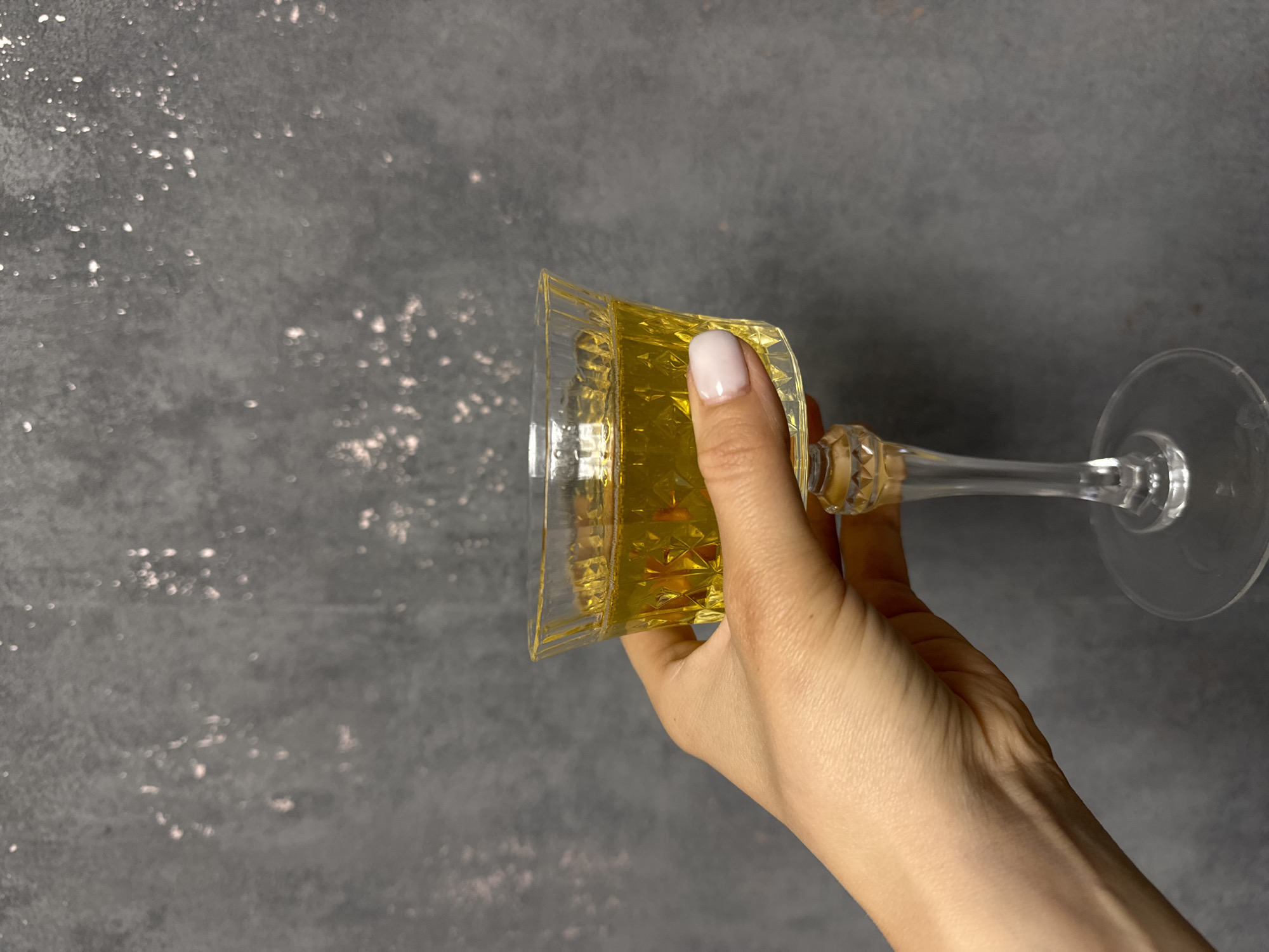 A hand with light skin and a pale pink nail holds a crystal glass filled with a golden liquid. The glass has a faceted bowl and a clear stem and base. The background is a dark gray, textured wall with some light pink speckles. The image is a close-up, focusing on the hand and the glass, with the background softly blurred. The scene appears to be indoors, but the specific location within Ponazyrevo, Russia, is not discernible from the visual information provided.