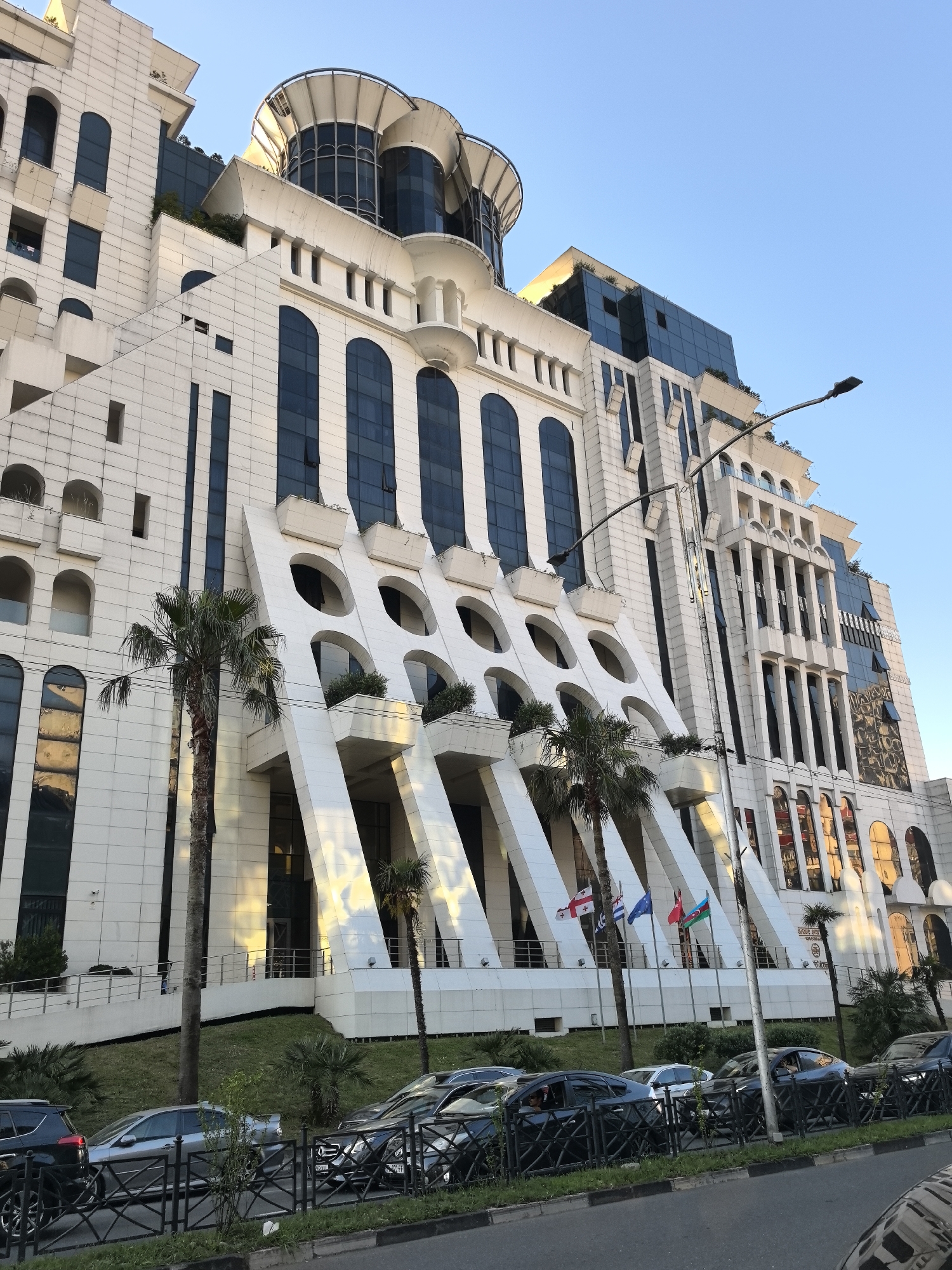 This is a medium-angle outdoor shot of a large, white, modern building with a distinctly geometric design. The building features numerous angular white supports, large arched windows, and glass-paneled sections. Palm trees are visible in front of the building, and several flags are planted in the ground. Cars are parked in a line along the street in front of the building, separated by a metal fence and a strip of grass. The sky is clear and blue, suggesting a sunny day. The lighting on the building indicates it is likely late afternoon or early evening, with the sun casting long shadows and highlighting some of the architectural features. There is no visible text in the image. The overall scene appears to be a well-maintained urban environment, possibly a hotel or a commercial complex.
