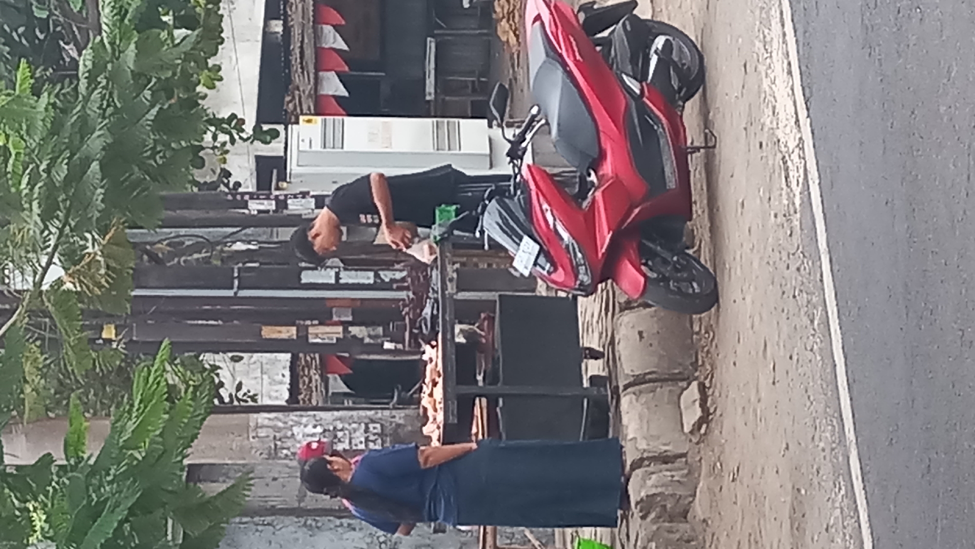 A street vendor is seen preparing food on a cart on the side of a road in Pare, Indonesia. The vendor, a man wearing a black t-shirt and a black cap, is seen handling food packaging. Beside him, a woman in a blue shirt and long blue skirt stands by the cart. The cart is laden with food items, appearing to be raw meat or poultry. A red scooter is parked in front of the cart. The background shows a building with a white facade and a red and white striped banner, possibly a storefront. Trees with green foliage are visible overhead. The ground is a mix of dirt and pavement, with a white line marking the edge of the road.