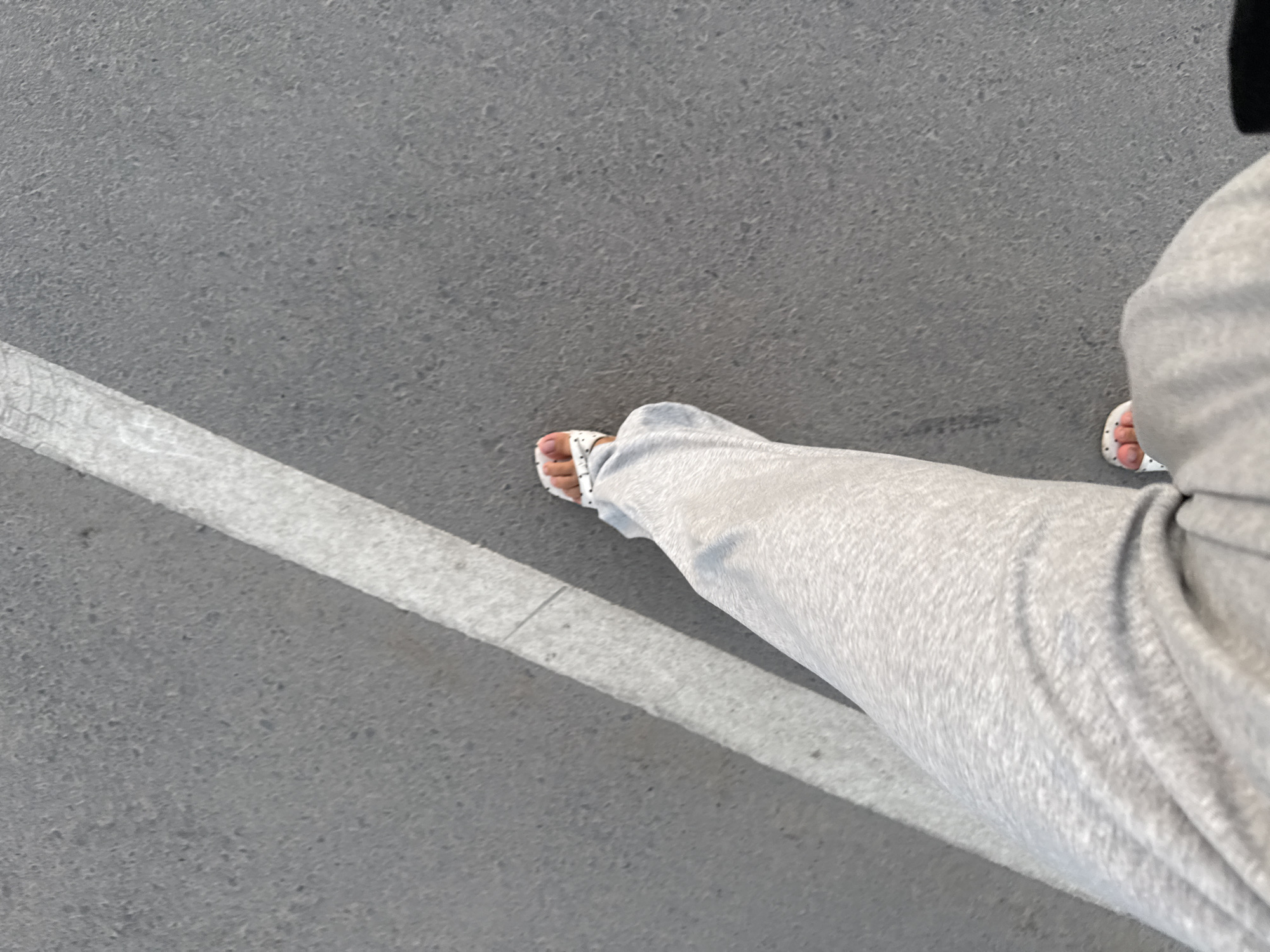 A low-angle, medium shot captures a person's legs and feet as they walk on a paved surface, likely a sidewalk or street in Bishkek City, Kyrgyzstan. A thick white line, possibly a road marking or boundary, runs diagonally across the frame from the upper left to the lower center. The pavement is grey and textured, with visible aggregate. The person is wearing loose-fitting light grey sweatpants and white sandals with a black polka-dot pattern. Their toes are visible in the sandals, and their right foot is slightly ahead of their left. The lighting suggests it is daytime, with a shadow cast by the white line onto the pavement, indicating the sun is positioned to the left of the frame. The scene is devoid of other people or significant urban features.