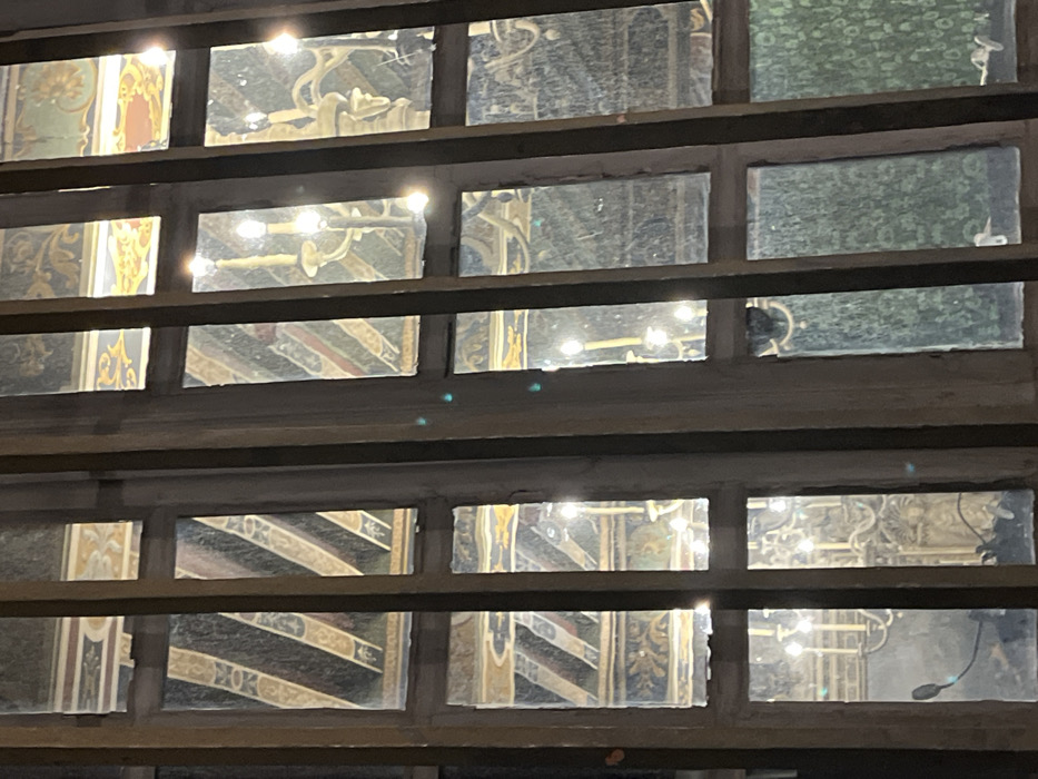 A multi-paned window offers a glimpse into a space adorned with ornate decorations. Chandeliers hang from the ceiling, reflecting light throughout the room. The walls feature intricate designs and patterns, suggesting a historical or religious setting. The view is partially obscured by the window frame, creating a layered perspective.