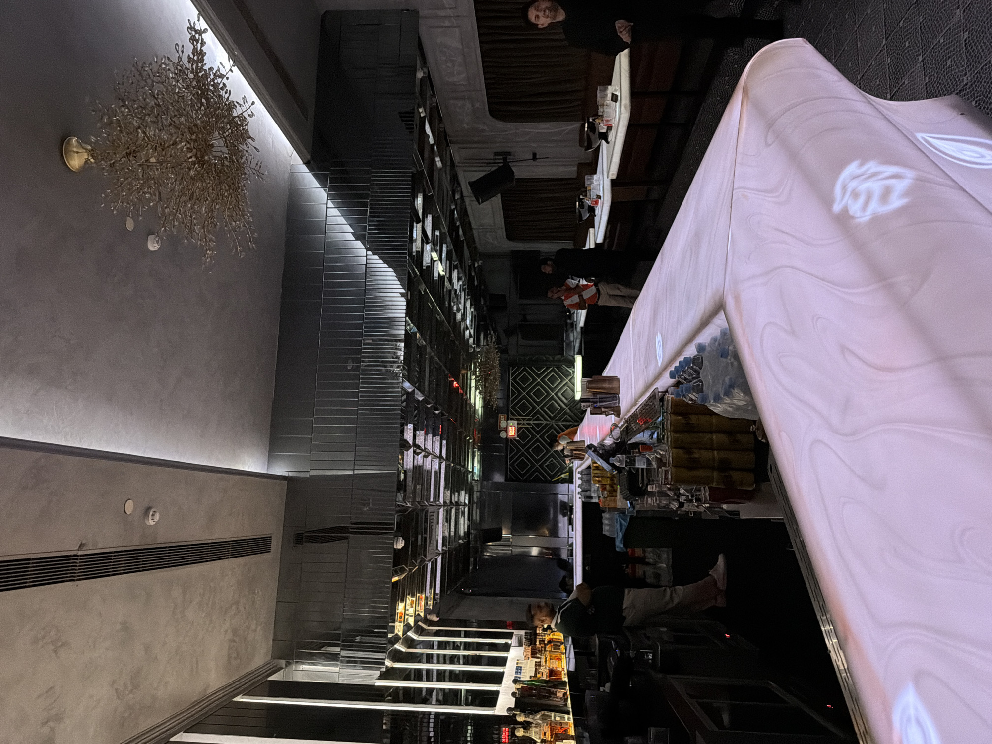 This image captures the interior of a modern, upscale bar or nightclub located in New Delhi, India. The perspective is from the front of a long, light-toned bar counter that extends from the lower left to the right side of the frame. The counter's surface exhibits a luminous, swirling pattern, possibly from a projection.
Behind the bar, a male bartender in a dark green polo shirt and light-colored pants stands looking towards the right, with various liquor bottles arranged on illuminated shelves behind him. The ceiling above the bar area features multiple reflective, geometric mirror panels, creating a fragmented visual effect. Vertical light strips and air conditioning vents are also integrated into the design. An "EXIT" sign is visible above a dark, patterned door in the mid-background on the left.
To the right of the bar, a seating area is visible with multiple tables and dark-upholstered booths against a wall with dark brown curtains. Several individuals, presumably staff members, are present in this area. One male staff member wearing an orange and white vest is visible in the mid-ground, while another man in a black shirt stands smiling towards the camera on the far right.
The ceiling throughout the venue is a light gray, textured surface, featuring recessed lighting, linear air vents, and smoke detectors. A prominent, ornate gold-colored chandelier, resembling a cluster of branches or foliage, hangs from the ceiling in the upper right quadrant of the image, contributing to the sophisticated ambiance. The overall lighting is dim and atmospheric, relying on artificial sources and reflections. The flooring appears dark and subtly textured.