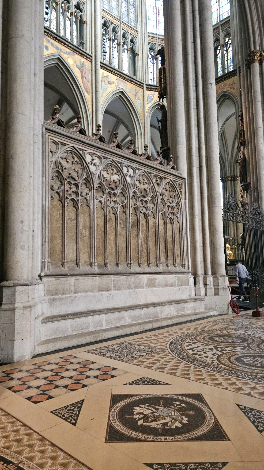 The image depicts the interior of a large Gothic cathedral, likely Cologne Cathedral, Germany, given the architectural style and historical context often associated with such grand structures. The scene focuses on the ornate detailing of the cathedral's architecture and interior.
In the foreground, a person, possibly a visitor or staff member, is visible near a wrought-iron gate. This individual is wearing a light-colored shirt and dark pants and appears to be pushing a red object on wheels, possibly a cleaning cart or part of a setup for an event. The floor is a complex mosaic pattern, featuring geometric shapes and circular inlays with intricate designs.
Dominating the mid-ground is a section of the cathedral wall adorned with a decorative stone screen featuring Gothic tracery. This screen is set against a textured, possibly gilded, background, suggesting it is a significant architectural or decorative element. Large, fluted stone columns flank this section, characteristic of Gothic cathedral construction.
In the background, the vaulted ceilings and stained-glass windows of the cathedral are visible, hinting at the immense scale and grandeur of the building. The overall impression is one of historical significance, architectural beauty, and the ongoing use of the space.