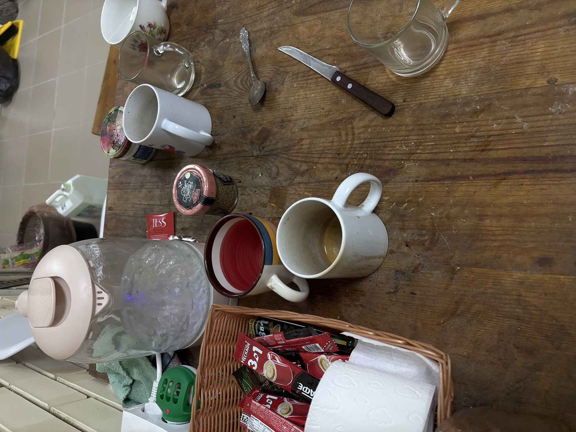 This image captures a forensic overhead view of a wooden table laden with items for hot beverage preparation in the city of Karasi, Russia. The table surface is worn, scarred, and exhibits multiple stains and debris, consistent with frequent, informal use.
The primary subject is a light pink and clear glass electric kettle, centrally positioned on the left, actively boiling water, indicated by visible bubbling and a subtle blue illumination from its base. Adjacent to the kettle, a green electronic insect repellent is plugged into a white wall outlet, next to a folded green cloth.
In the lower left, a woven light brown basket contains an assortment of single-serving instant coffee packets, including brands such as "MECCADIM 3в1," "НЕСКАФЕ 3в1," and "MONARCH 3в1," alongside "CEYLON TEA" packets. White paper towels, embossed with a leaf pattern, partially cover the contents of the basket.
Several mugs and glasses are distributed across the table:
* A ceramic mug with horizontal red, blue, yellow, and brown stripes contains a reddish-brown liquid.
* A plain white ceramic mug, positioned below the striped mug, is largely empty but heavily stained with dried brown liquid residue both inside and on its exterior.
* A clear glass mug, empty, is situated to the right of the stained white mug.
* A white ceramic mug with a red floral pattern near its rim contains a white tea bag tag, suggesting recent tea consumption.
* Another clear glass mug, empty, is located in the lower right quadrant of the table.
Two jars are visible behind the mugs: one with a pink lid, labeled "Chef's Blends," containing a granular brown substance, and another with a multicolored floral lid, contents unidentifiable. A small, ornate silver spoon and a utility knife with a dark wooden handle are also on the table surface.
The background shows a partially visible interior, including white horizontal wall panels on the left, and a tiled floor, a dark plastic bag, a brown basket, and a white plastic container (jerrycan) further back on the right. No individuals are present in the scene. The arrangement of objects suggests an area dedicated to casual beverage preparation and consumption.