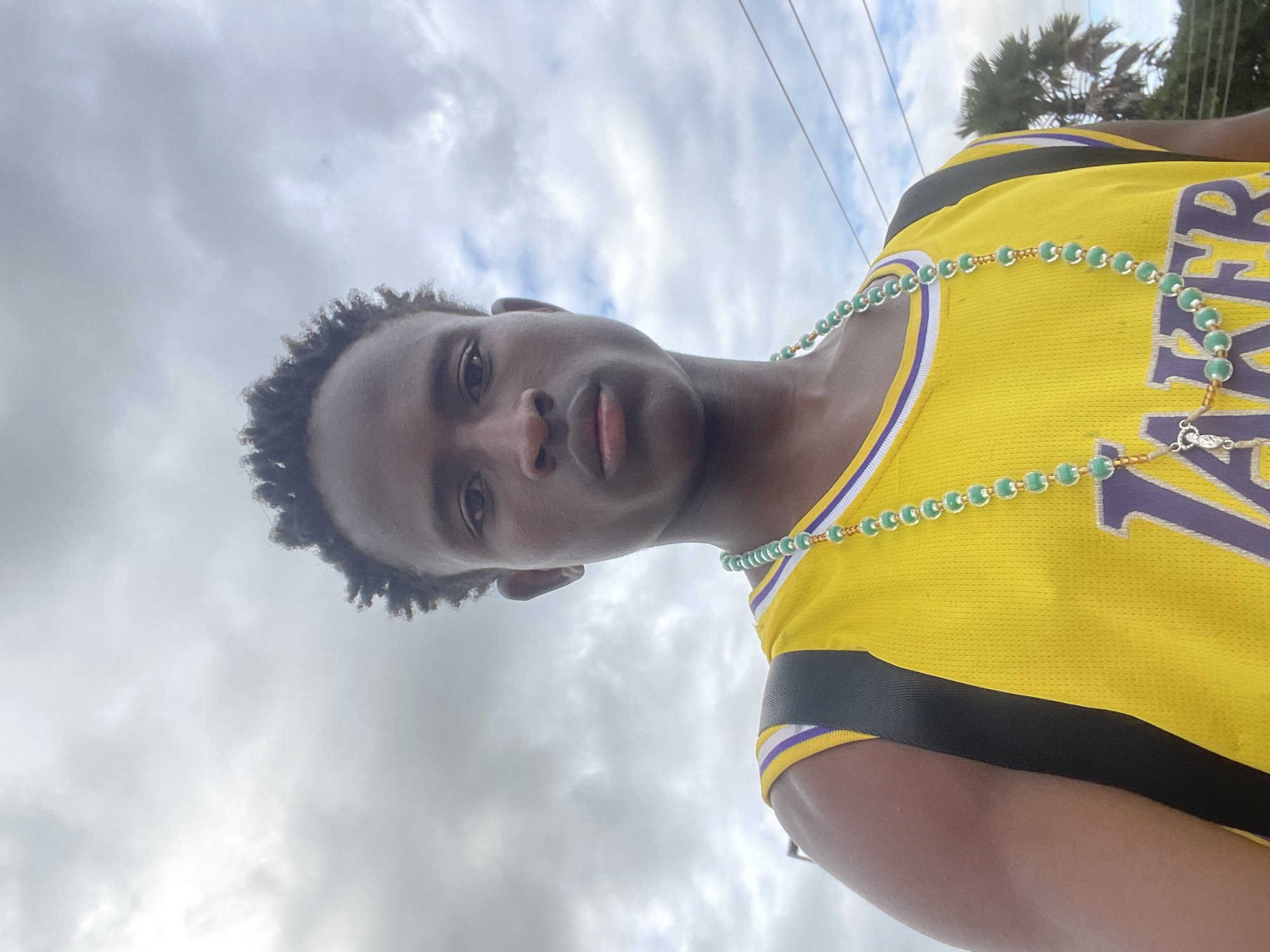 A medium shot shows a young Black male with short, dark, curly hair, wearing a yellow basketball jersey with purple trim and lettering. He is also wearing a black backpack and a beaded necklace with green and gold beads, featuring a small silver charm at the bottom. The man is looking directly at the camera with a neutral expression. The background consists of a cloudy sky and the tops of palm trees, suggesting an outdoor setting. The overall lighting is diffuse, indicating an overcast day. The specific location in Jos, Nigeria, cannot be determined from the visual information in the image.