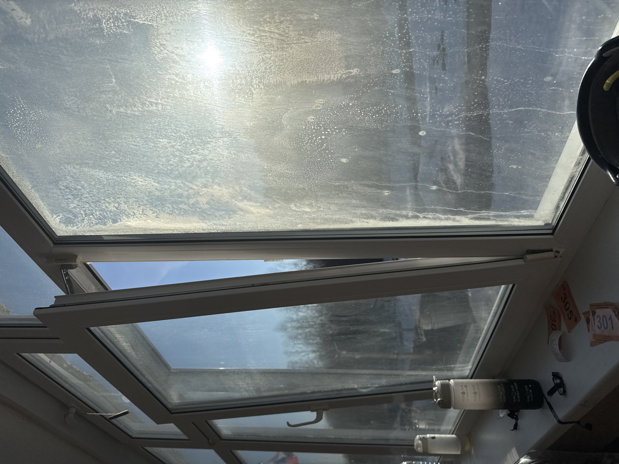 The image depicts a view looking up through a glass roof, possibly a conservatory or greenhouse structure. The glass panels are streaked and dirty. One panel is open, revealing a view of the sky and trees outside. On the interior frame, there are objects including a water bottle and what appear to be numbered tags or labels. The specific location in Usolye, Russia cannot be determined from this image. There are no people visible.