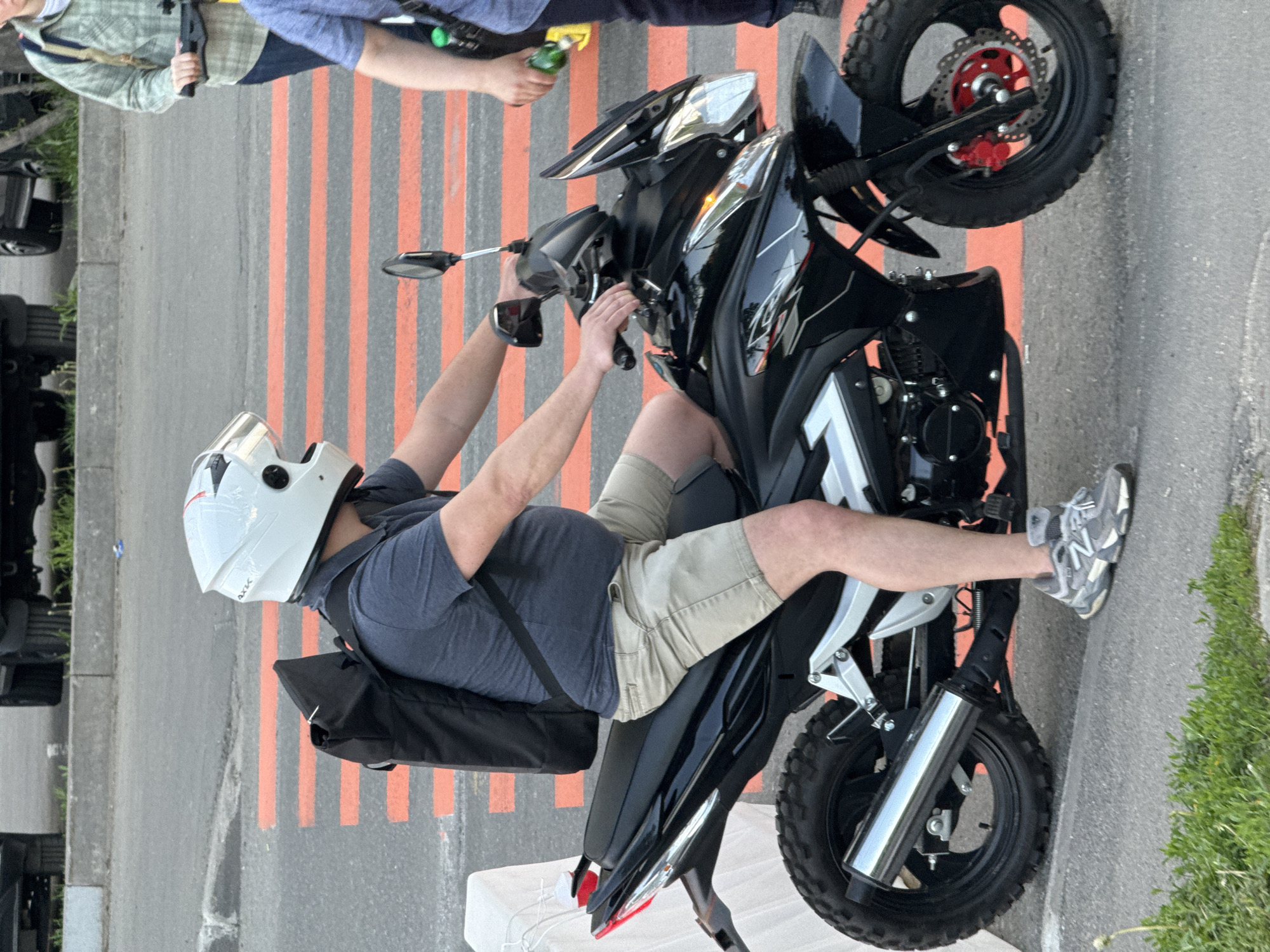 A man wearing a white helmet, a blue shirt, and khaki shorts sits astride a black and white motorcycle on a street with orange and gray striped pedestrian crosswalk markings. He has a black bag strapped across his back. The motorcycle has knobby tires, and the front wheel is turned slightly to the left. Other individuals and vehicles are visible in the background. The location is specified to be Moscow, Russia.