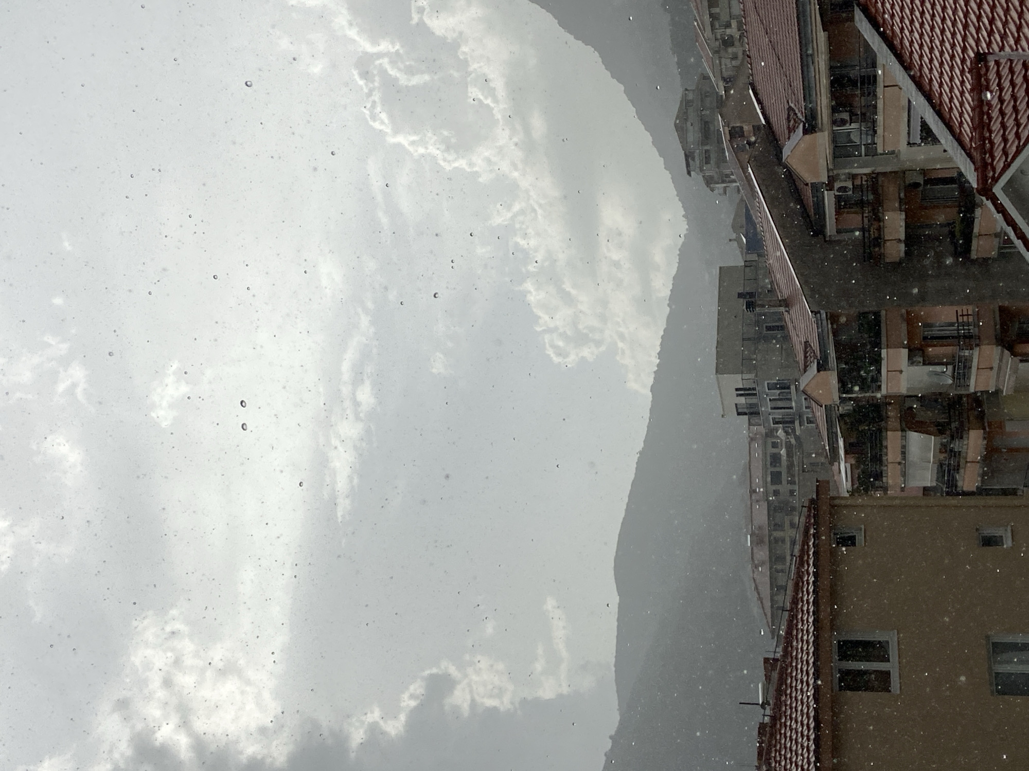 The image captures a view of Budva, Montenegro, during a rainfall. Water droplets are visible on the foreground, suggesting the photo was taken from a window or similar vantage point. In the middle ground, residential buildings with red-tiled roofs are clustered together, with some featuring balconies and air conditioning units. Further back, more buildings and distant, hazy mountains can be seen, their forms softened by the atmospheric conditions. The sky is overcast with dramatic clouds, and the overall lighting suggests a gloomy, rainy day. No people or specific actions are discernible in the scene.