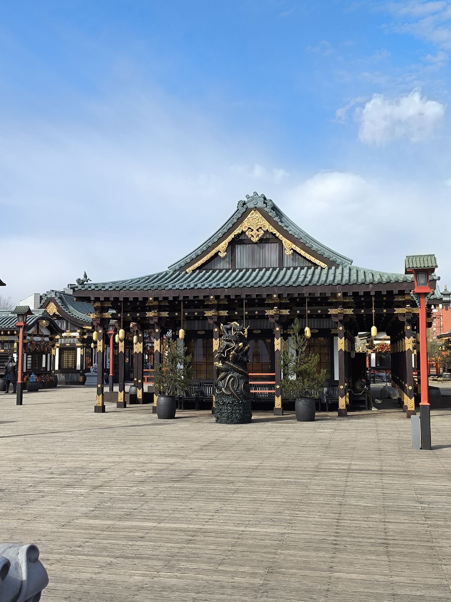 The image depicts a Japanese-style pavilion in a public space. The pavilion has a multi-tiered roof with dark green tiles and gold accents. It is supported by dark wooden pillars adorned with gold details. Potted trees are positioned near the pavilion's entrance. A sculpted dragon statue stands prominently in front of the building. The pavilion is situated on a wooden deck, which extends into an open area. In the background, other similar pavilions and street lamps can be observed. The sky is blue with scattered clouds. This location is specifically identified as the "Island of Dreams" amusement park in Moscow, Russia. In one of the other buildings, a person is sitting.