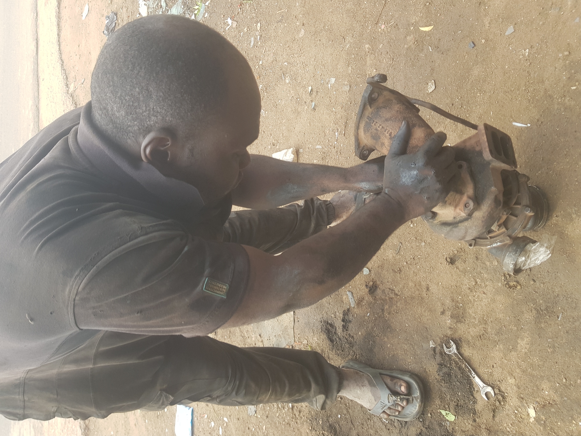 In Kufena, Nigeria, an adult Black male is kneeling on a dusty, bare ground surface, examining a large, dirty, metallic, mechanical component. His clothing consists of a dark, likely black or grey, collared shirt, and pants of a similar dark color. His hands are covered in dark grime. He is holding and manipulating the component with both hands. The ground also has a wrench nearby, as well as a plastic bag.