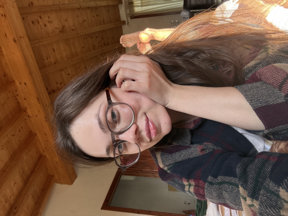 A young adult female is depicted in a medium shot indoors, within an unidentifiable structure located in Monsummano Terme, Italy. She is positioned towards the camera, smiling. She wears round spectacles with a translucent grey frame and a multi-colored plaid shirt featuring dark blue, maroon, beige, and green tones. Her right hand supports the right side of her face. Her long brown hair frames her face and extends over her shoulders. In the background to the left, a portion of a wooden door within a light-colored frame is visible. To the right, two bare feet are propped upwards, strongly illuminated by a natural light source. A dark, rectangular object, possibly luggage, is partially visible near the feet. The ceiling overhead is composed of visible wooden beams and planking.
