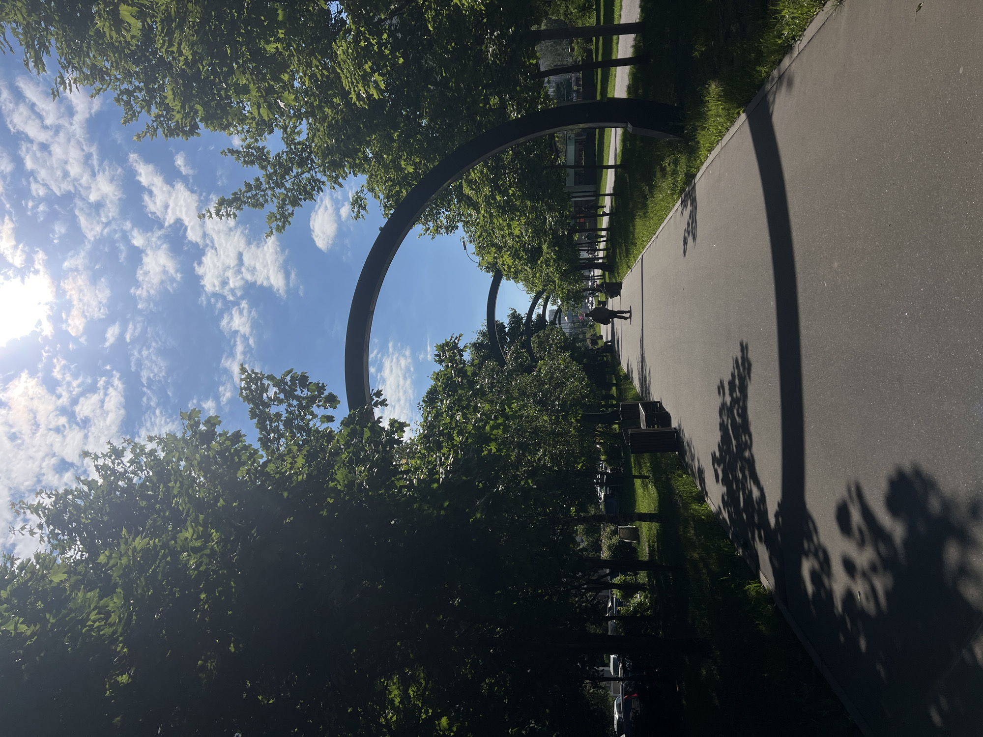 This image captures a wide-angle view of a modern park or promenade in Moscow, Russia, on a bright, sunny day. A broad, paved pathway, likely asphalt, extends into the distance on the right side of the frame, bordered by neatly maintained grass and dense green trees on both sides.
Several large, dark, metallic arching structures are a prominent feature, spanning over the pathway at regular intervals, suggesting a decorative or architectural installation. These arches and the trees cast long, distinct shadows across the pathway, indicating strong overhead sunlight. The sun itself is visible as a bright flare in the upper left corner, illuminating a clear blue sky adorned with scattered white, puffy clouds.
In the middle distance, a solitary person is walking away from the viewer along the pathway. Along the left side of the pathway, beneath the canopy of trees, several park benches are visible, and further back, what appear to be parked vehicles can be discerned, hinting at an urban setting surrounding the park. The scene conveys a sense of peaceful leisure and pleasant outdoor activity under excellent weather conditions. No visible text is present in the image.