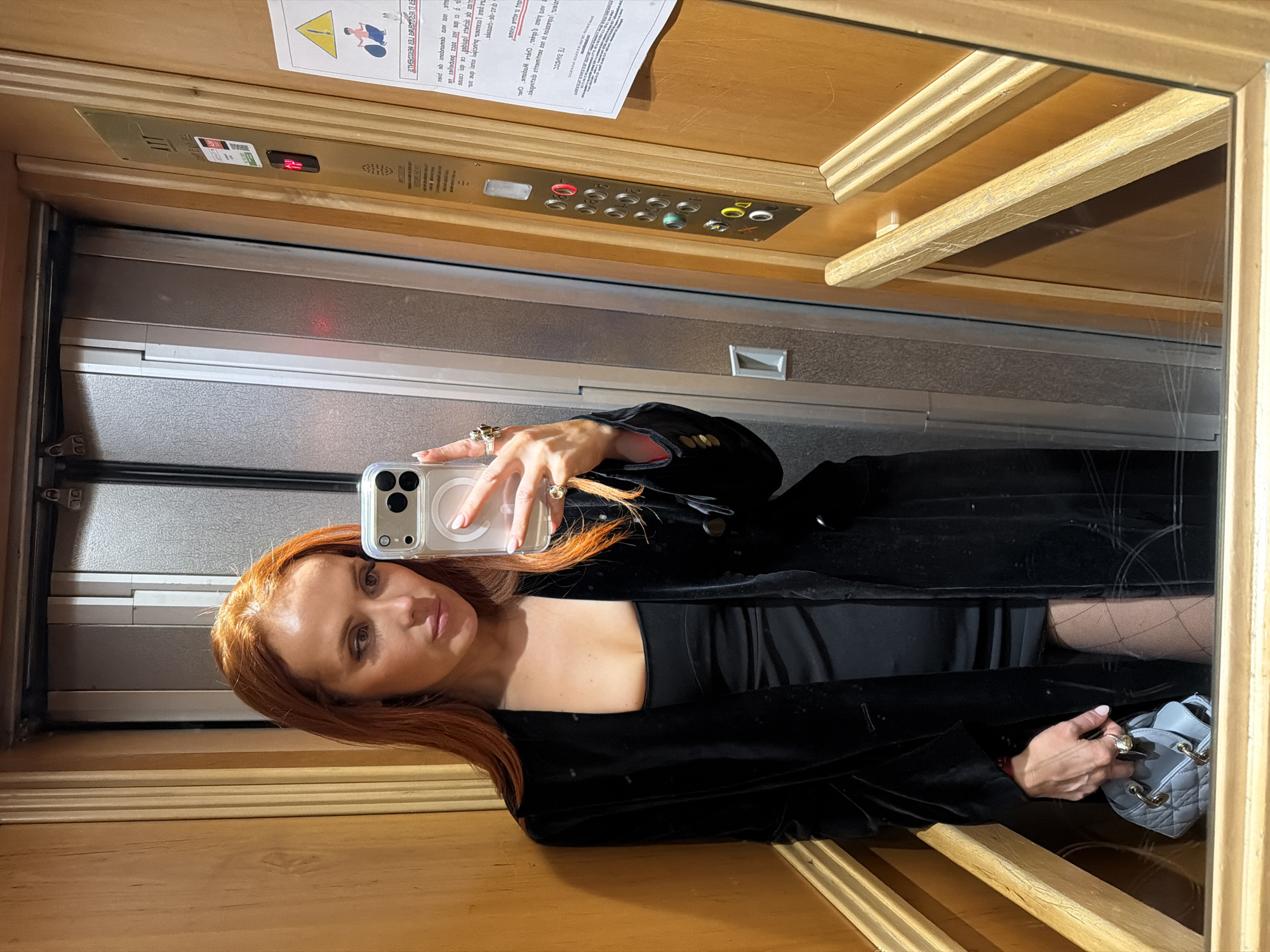 A woman with long red hair takes a selfie in a mirrored elevator. She wears a black dress and a black velvet jacket with gold buttons. She holds a blue purse in her right hand and a clear phone case in her left. The elevator has wood paneling with gold trim, and silver doors. Elevator buttons are visible above the woman's head. A warning sign written in a foreign language is posted on the wall above the elevator buttons. The woman is wearing a large gold ring.