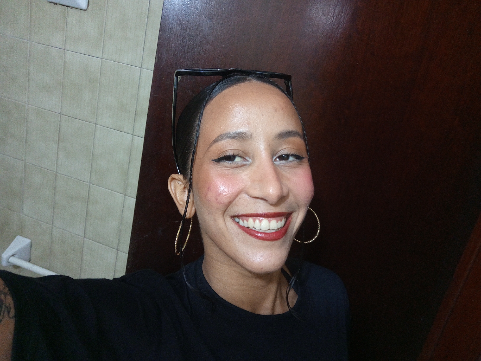 A medium-complexion woman with dark hair pulled back and adorned with eyeglasses on top of her head smiles at the camera. She is wearing large hoop earrings and dark lipstick. The setting appears to be indoors, with tiled walls to the left and a dark, wooden-paneled wall on the right. It is not possible to determine the location in the city of Porlamar, Venezuela, from the image alone.