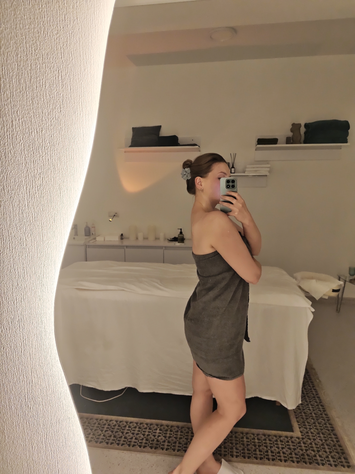 A young woman, seen from the side and reflected in a large, wavy mirror, stands in a room that appears to be a spa or massage treatment room. She is wearing a dark grey towel wrapped around her body. She holds a mobile phone in her right hand, taking a selfie. Her hair is pulled back in a bun with a grey clip.
The room has a light-colored, textured wall to the left, illuminated by an unseen light source. In the mirror's reflection, a massage table covered in white sheets is visible. Behind the table, a counter with various bottles and containers suggests a treatment area. Shelves on the wall above the counter display dark-colored pillows, towels, and decorative items like a small bear figurine. The overall lighting in the room is warm and subdued, suggesting it might be evening or a low-light environment. The floor has a patterned rug. The location is identified as Vinnytsia, Ukraine, but no specific text is visible in the image to confirm this. The woman's pose and the setting suggest a moment of preparation or relaxation.