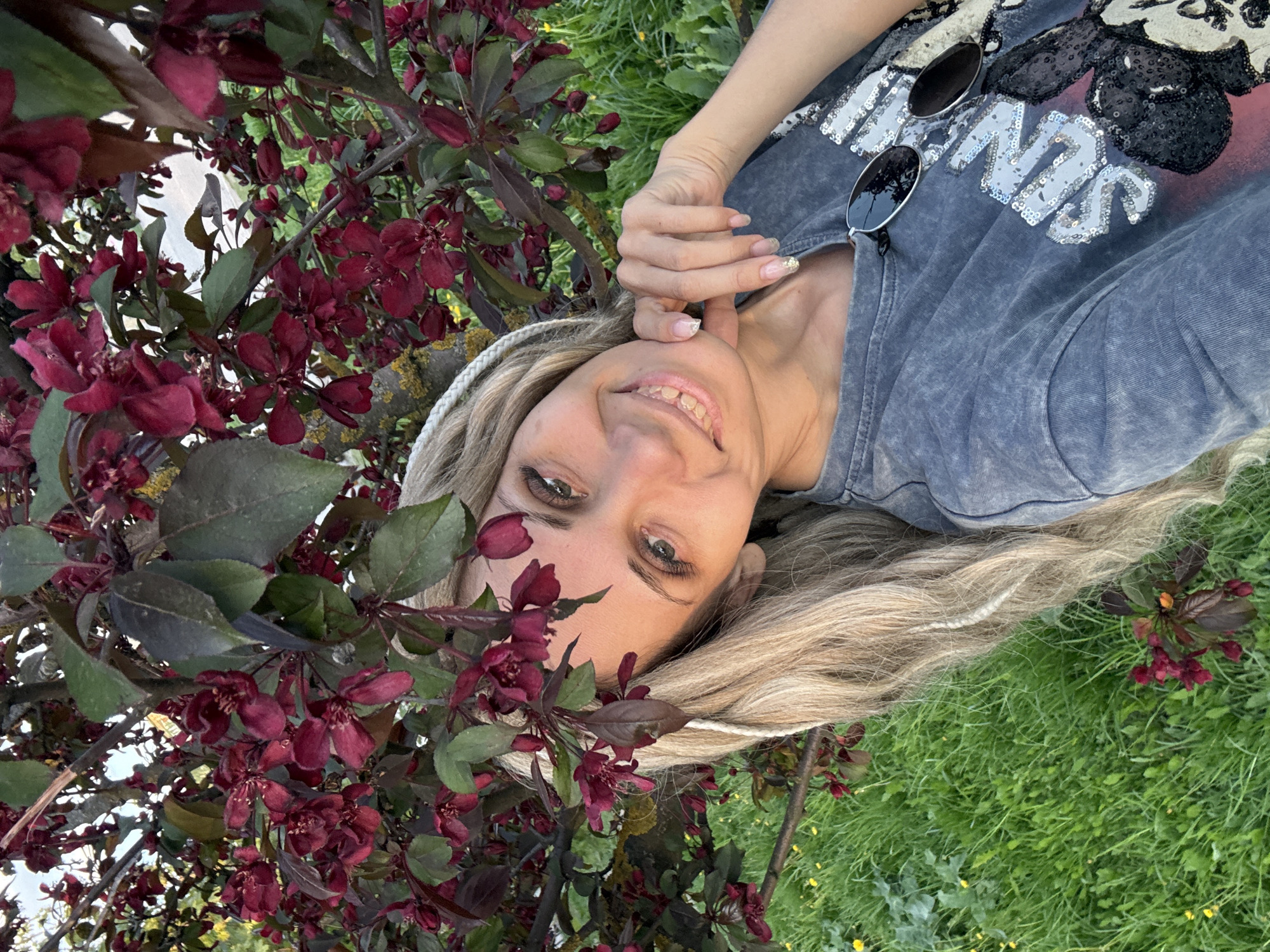 A woman is lying on the grass next to a blossoming tree in Rusanovo, Russia. The tree has dark red leaves and flowers. The woman is fair-skinned with blonde hair and green eyes. She is wearing a gray t-shirt with sunglasses on her chest. She is smiling and looking at the camera, with her right hand touching her chin. The grass is green and there are small yellow flowers scattered throughout.