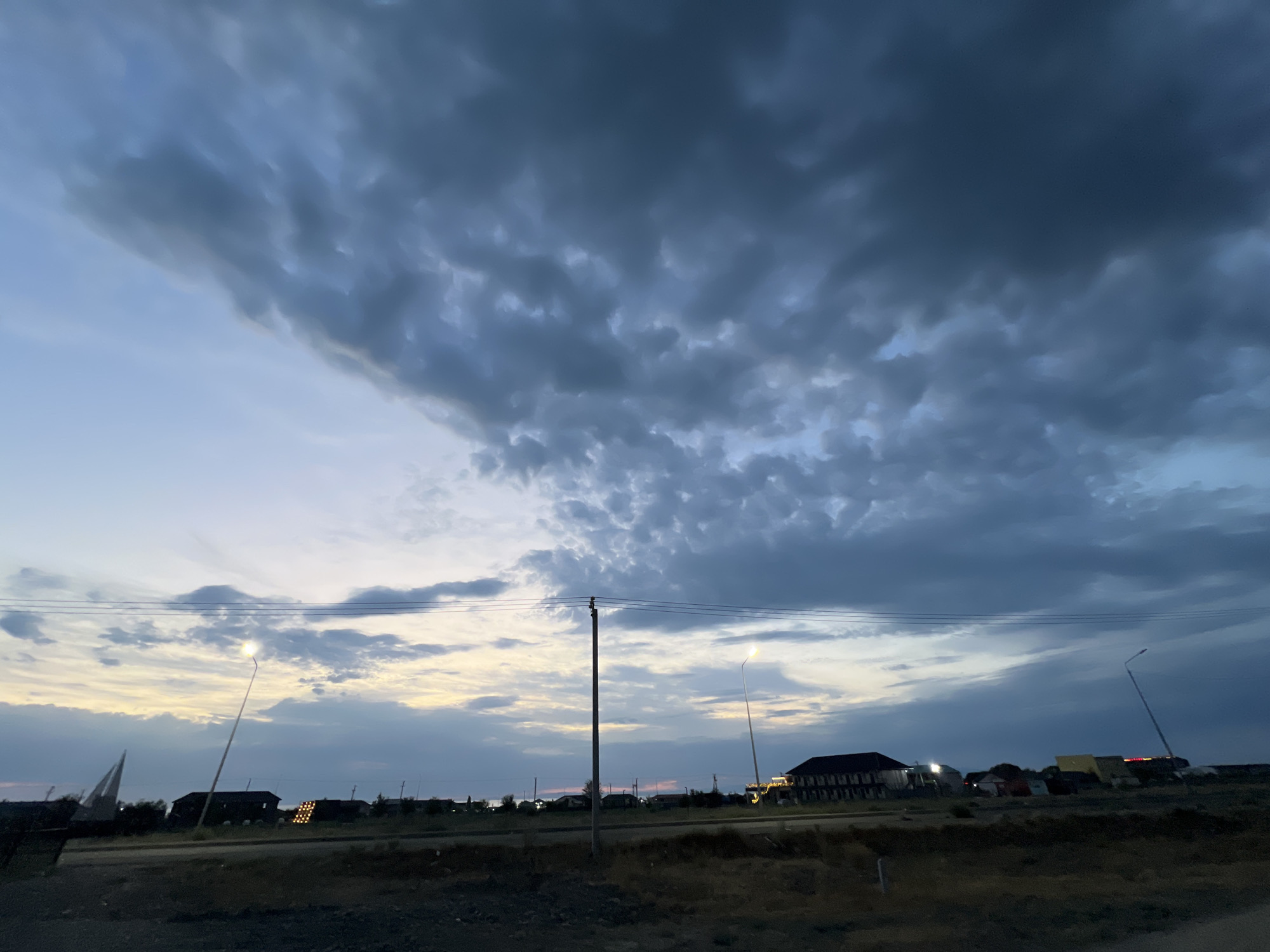 The image captures a twilight scene in the city of Ақши, Kazakhstan. A dark, heavily clouded sky dominates the upper portion of the frame, with lighter hues of blue and yellow visible near the horizon. Several buildings, including a large, multi-story structure with illuminated windows, are scattered across the landscape. Electrical poles with overhead wires and streetlights are present, with at least two lights actively illuminated. A partially visible, triangular structure, possibly a sail or a wind sculpture, is situated on the left side of the image. The ground in the foreground appears to be rough terrain, possibly a dirt road or undeveloped land. The overall atmosphere suggests either the end of a day or the approach of a storm.