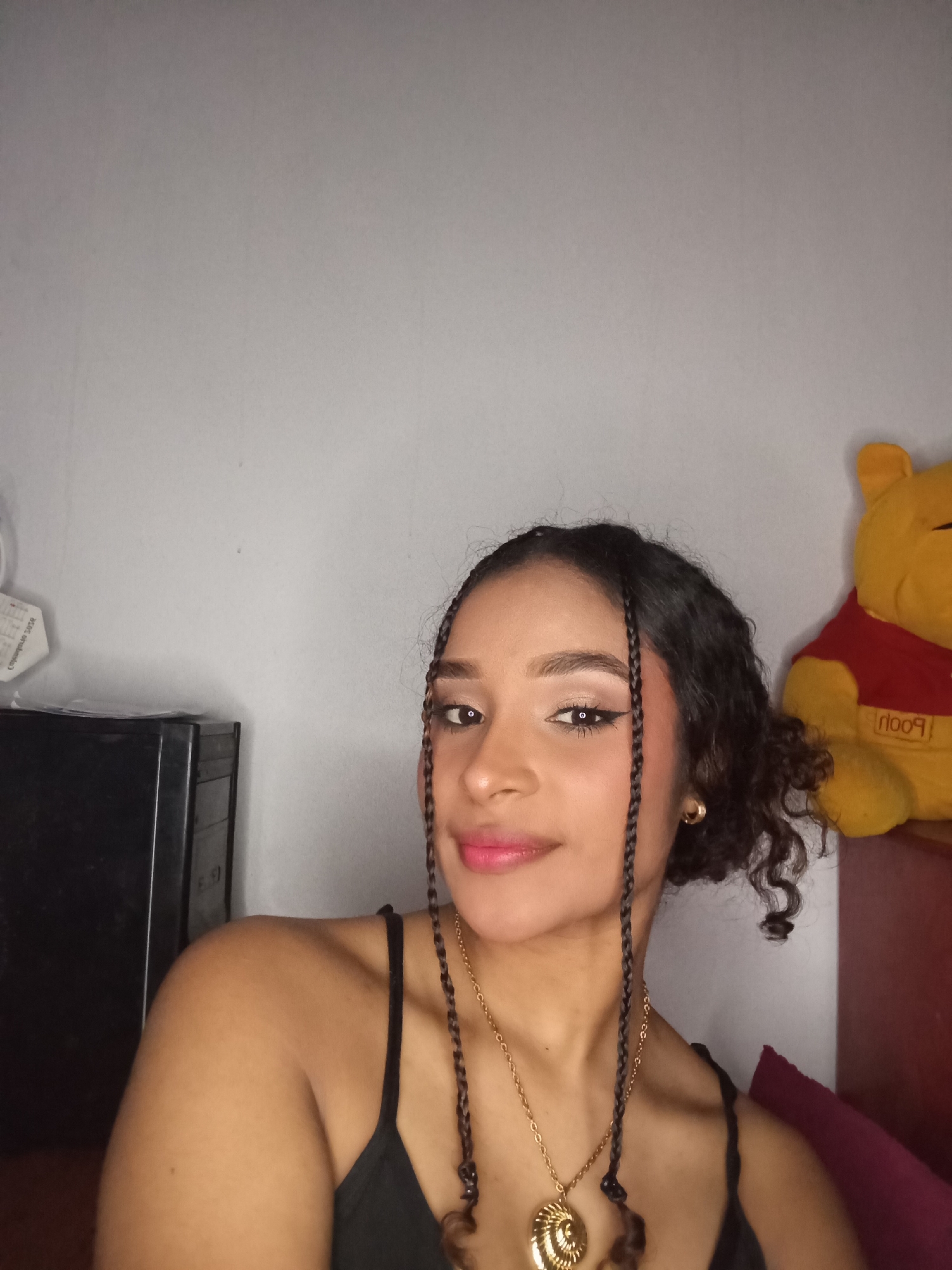 The scene is an indoor setting in Juan Griego, Venezuela, featuring a portrait of a female individual. The subject is a young woman with a medium-dark complexion, dark curly hair, and several small braids framing her face. She is looking directly at the viewer with a slight smile. She wears a black spaghetti-strap top, a gold-colored chain necklace with a large, ornate, spiral-shaped pendant, and a small gold hoop earring on her visible right ear. Her face shows visible makeup, including winged eyeliner, light eyeshadow, and pink lipstick. The background consists of a plain, light grey wall. To the subject's left, a dark rectangular object is partially visible, with a crumpled white paper item displaying partially legible text ("comprabado 2024" or similar) affixed to the wall above it. To the subject's right, a yellow stuffed Winnie the Pooh bear with a red shirt (text "POOH" visible, possibly inverted) rests on a dark reddish-brown surface, partially obscuring a dark red cushion or pillow.