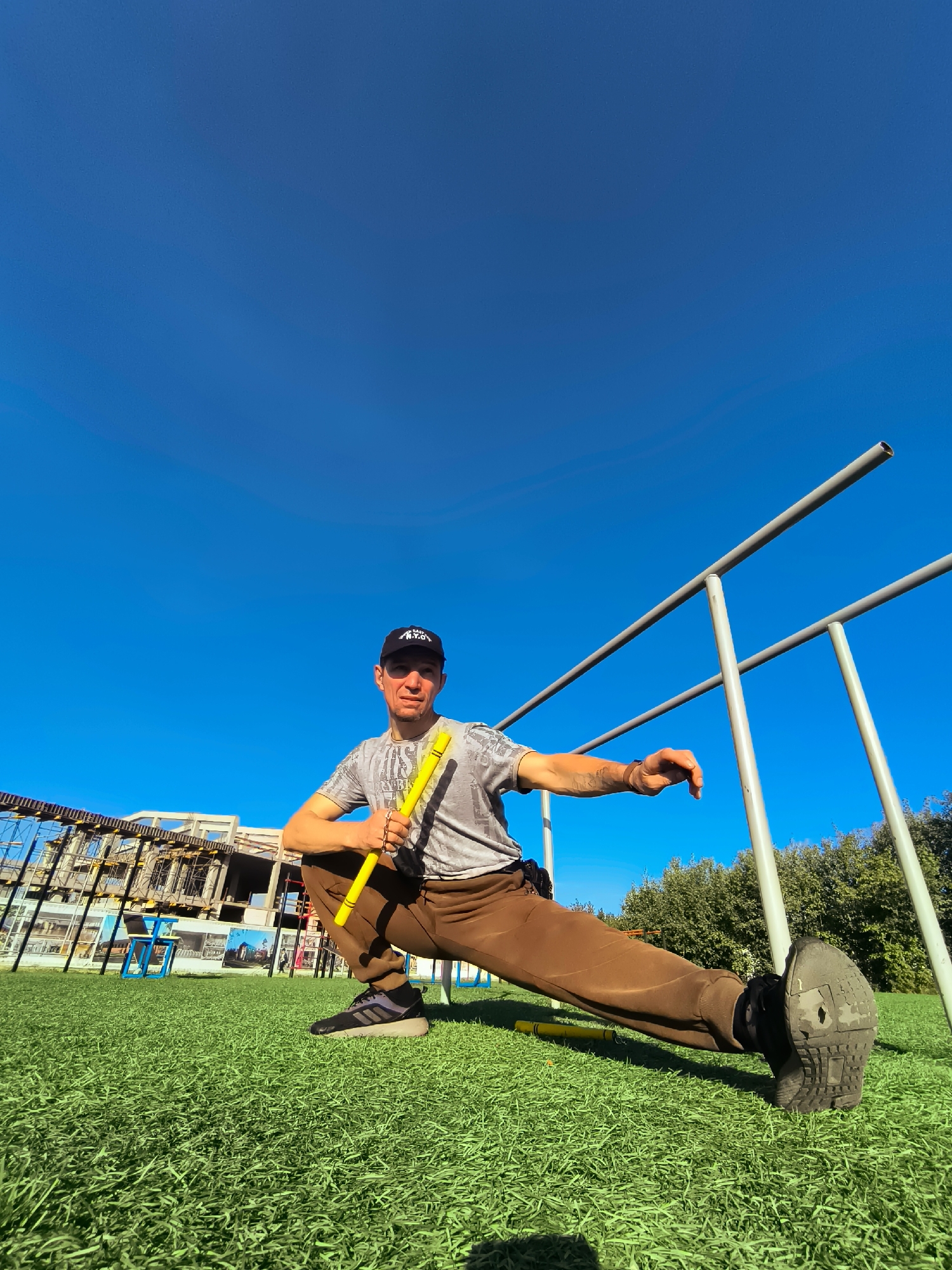 A man, wearing a grey t-shirt, brown sweatpants, a black baseball cap, and black athletic shoes, is performing a leg stretch outdoors on a bright, sunny day. He is holding a yellow stick with both hands and his left leg is extended forward, with his right leg bent underneath him. The ground is covered in bright green artificial turf. In the background, there are metal parallel bars, trees, a blue chair, and a building under construction. The sky is a clear, vibrant blue. The image appears to be taken in the city administration area of Qarağandı, Kazakhstan, likely at an outdoor sports or recreational facility.