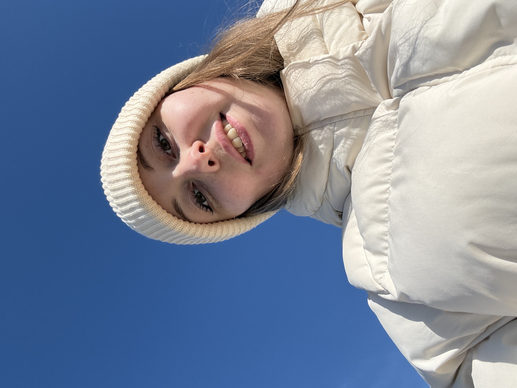 A woman wearing a white knit hat and white puffy jacket is seen from a low angle, against a clear blue sky. The location is Unknown, Unknown. She is smiling slightly. The sunlight is casting shadows on her jacket.