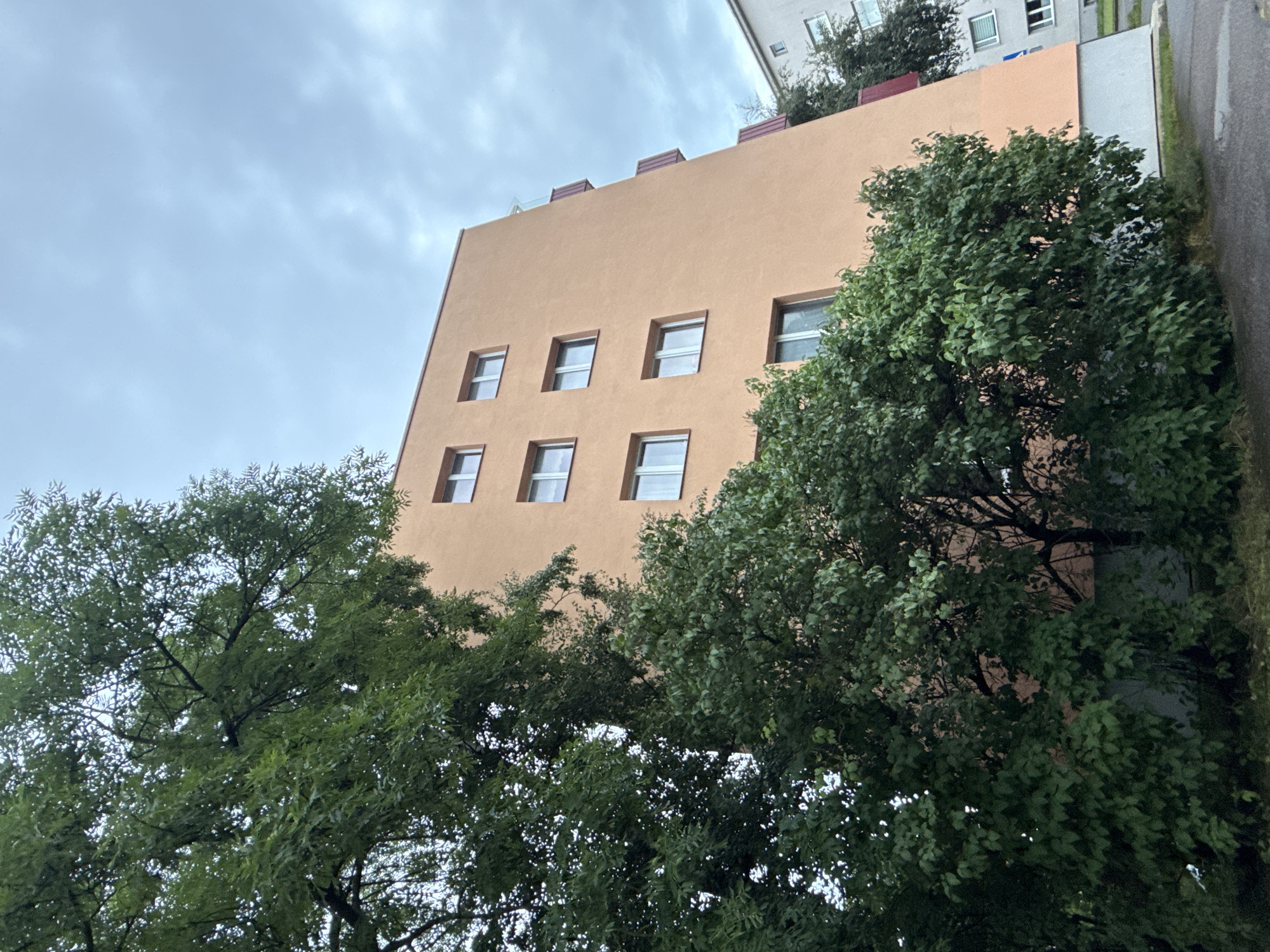 The image depicts an exterior view of residential buildings in Tallinn, Estonia, under a cloudy sky. In the foreground, lush green trees partially obscure a multi-story apartment building with a peach-colored facade. The building features a series of rectangular windows arranged in horizontal rows. Red balconies with vertical supports are visible on the right side of the peach-colored building, extending downwards. Adjacent to it, a lighter-colored building, possibly gray or white, is also visible on the right, with its own set of windows. The ground in the lower portion of the image appears to be a paved surface, possibly a sidewalk or road, with some wet patches suggesting recent rain. The overall scene conveys an urban residential environment with significant vegetation.