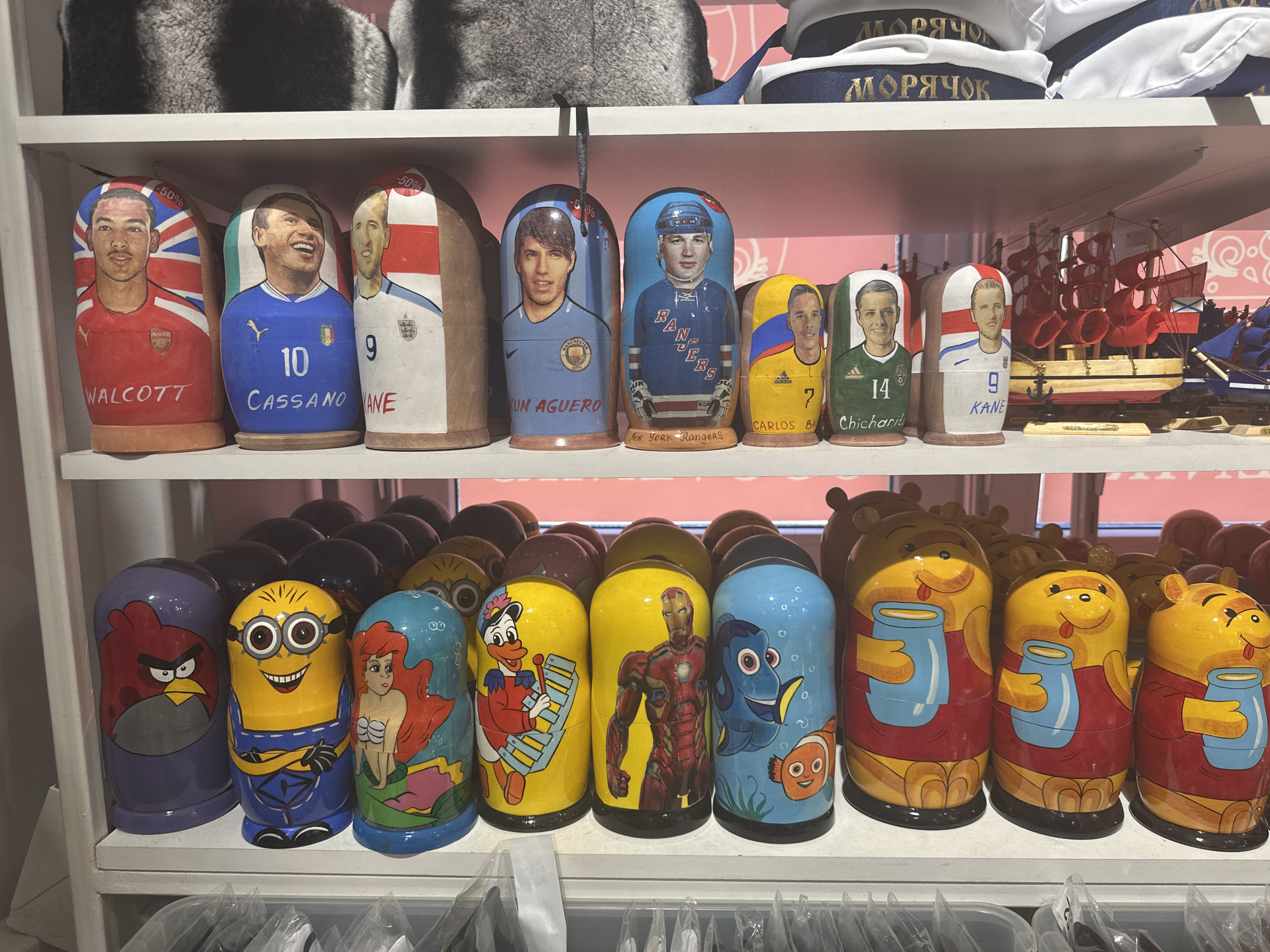 The image depicts two shelves filled with Russian nesting dolls, likely in Saint Petersburg, Russia. The top shelf features dolls with portraits of soccer and hockey players, including Walcott, Cassano, Agüero, and Kane. The bottom shelf displays dolls decorated with cartoon characters like Angry Birds, Minions, Ariel, Donald Duck, Iron Man, Nemo, and Winnie-the-Pooh. Above the top shelf is a fur hat and a sailor hat. Below the bottom shelf are items covered with plastic.